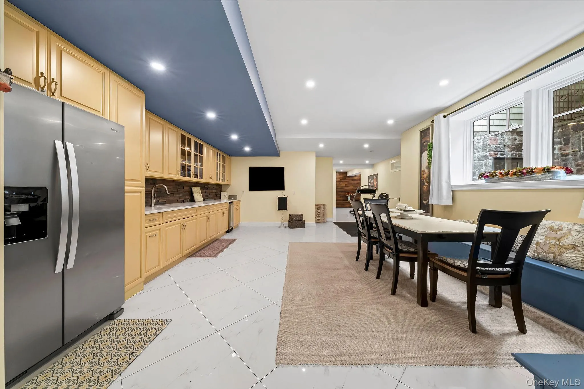 Kitchen featuring stainless steel refrigerator, light wood cabinetry, stone-texture backsplash, recessed lighting, and large-format tile flooring Kitchen featuring stainless steel refrigerator, light wood cabinetry, stone-texture backsplash, recessed lighting, and large-format tile flooring