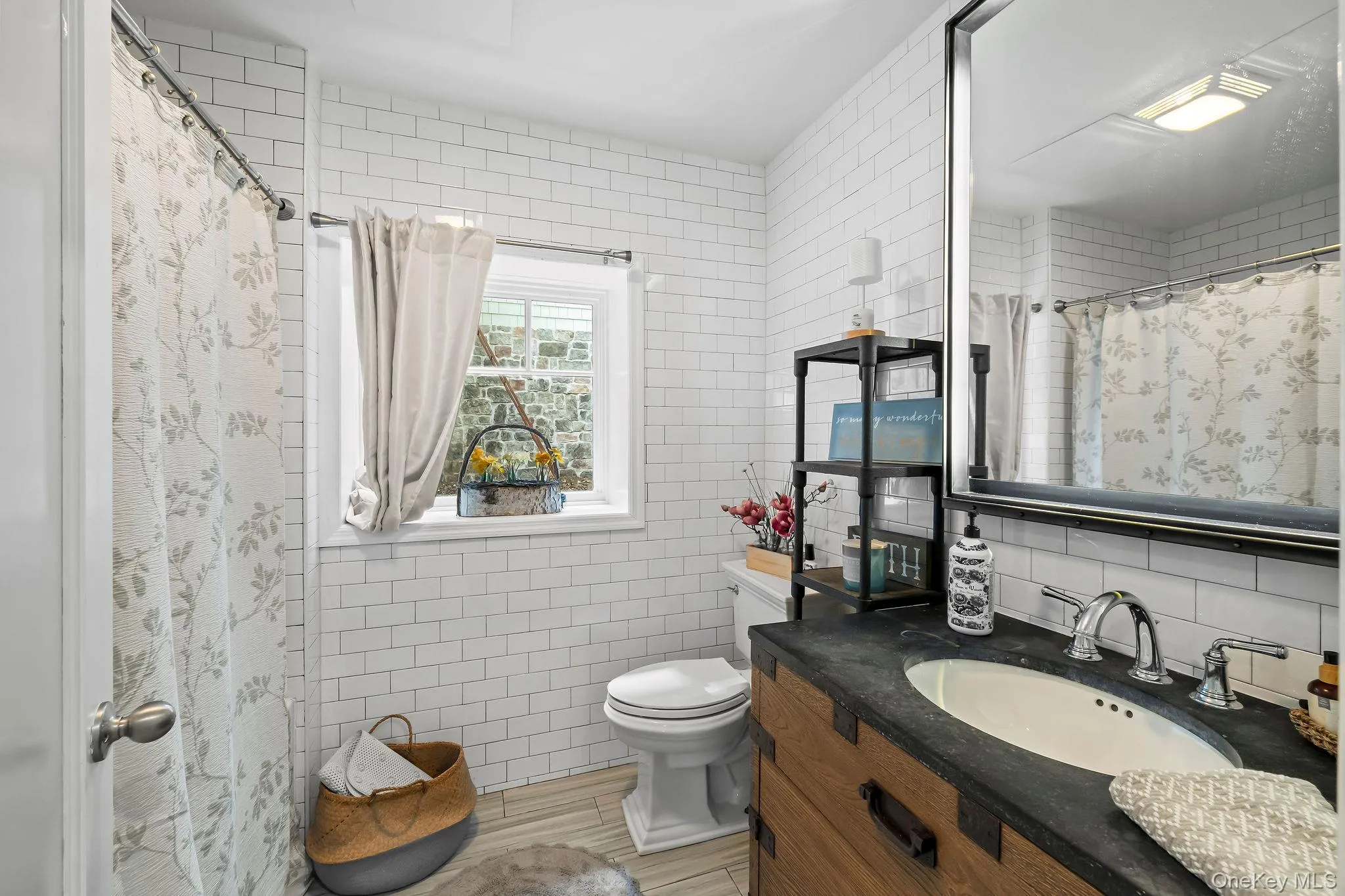 Bathroom featuring white subway tile wall cladding, wood-finish vanity with a dark countertop and integrated sink, large-format framed mirror, and wood-finish flooring Bathroom featuring white subway tile wall cladding, wood-finish vanity with a dark countertop and integrated sink, large-format framed mirror, and wood-finish flooring