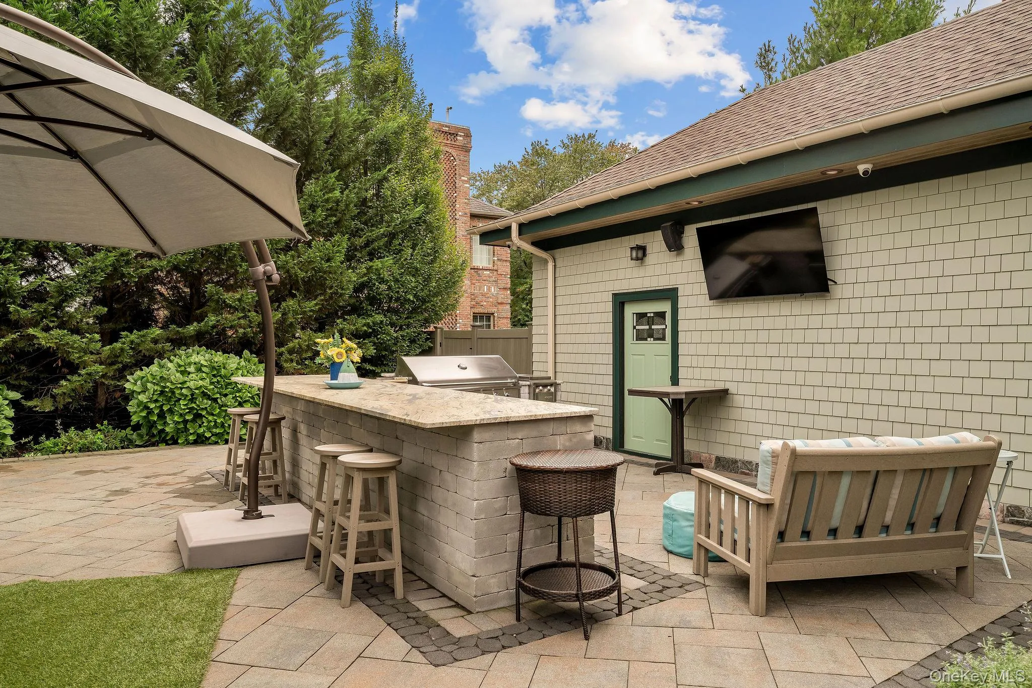 Outdoor patio featuring a stone-paver hardscape, built-in masonry grill island with a stone countertop, and exterior wall-mounted television Outdoor patio featuring a stone-paver hardscape, built-in masonry grill island with a stone countertop, and exterior wall-mounted television