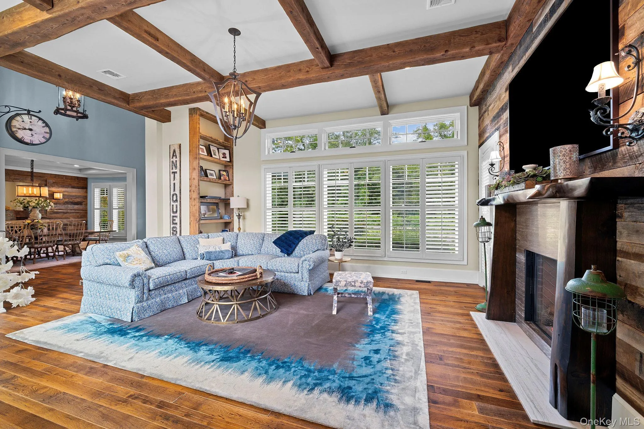 Living area featuring exposed wood ceiling beams, wood-finish flooring, a floor-to-ceiling wood plank accent wall, a fireplace with a live-edge wood mantel, and windows outfitted with plantation shutters Living area featuring exposed wood ceiling beams, wood-finish flooring, a floor-to-ceiling wood plank accent wall, a fireplace with a live-edge wood mantel, and windows outfitted with plantation shutters