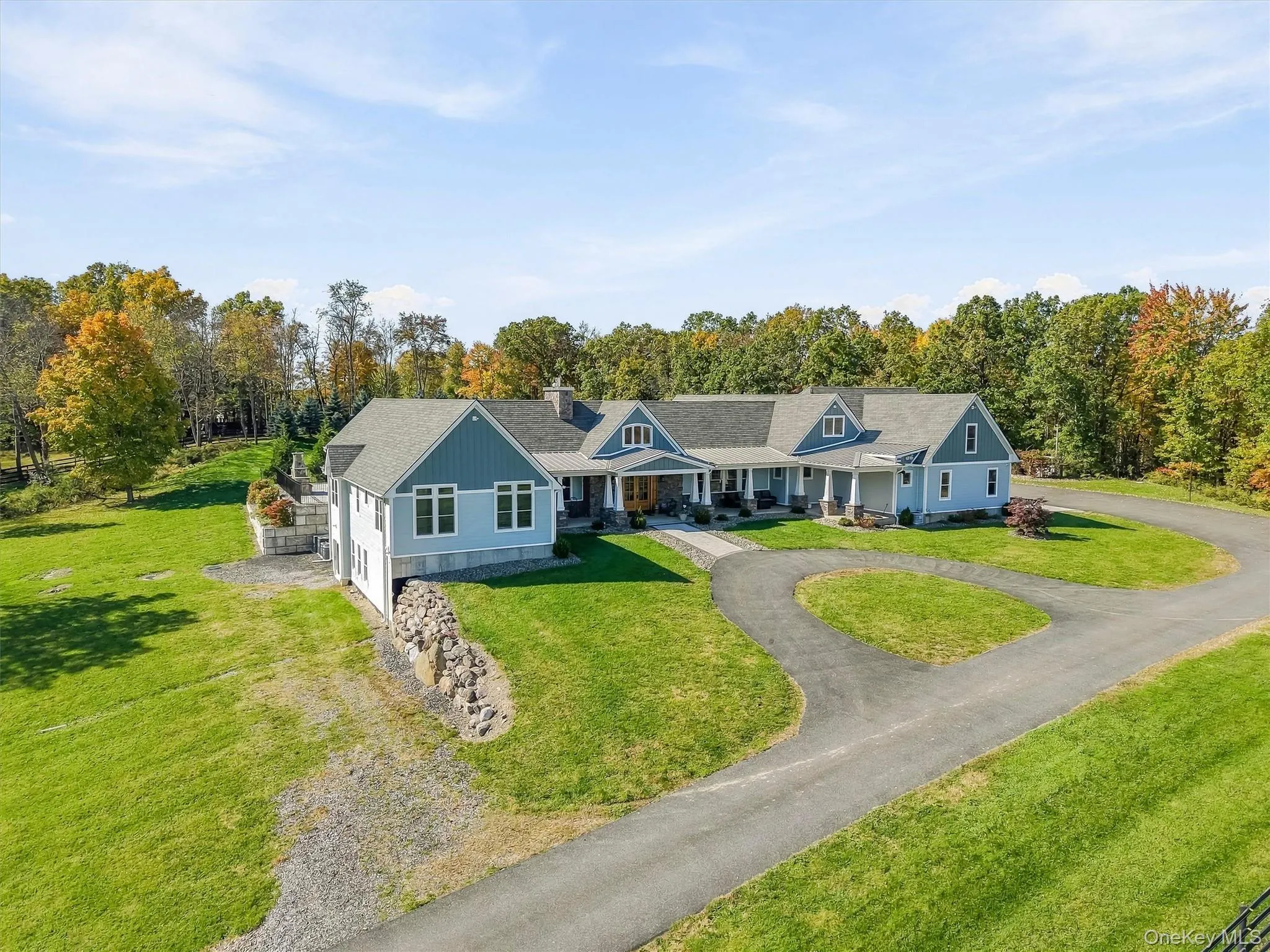 Expansive residence featuring a multi-gabled roofline, lap siding exterior, a wrap-around covered porch with stone-base columns, a circular driveway, and tiered stone retaining walls Expansive residence featuring a multi-gabled roofline, lap siding exterior, a wrap-around covered porch with stone-base columns, a circular driveway, and tiered stone retaining walls