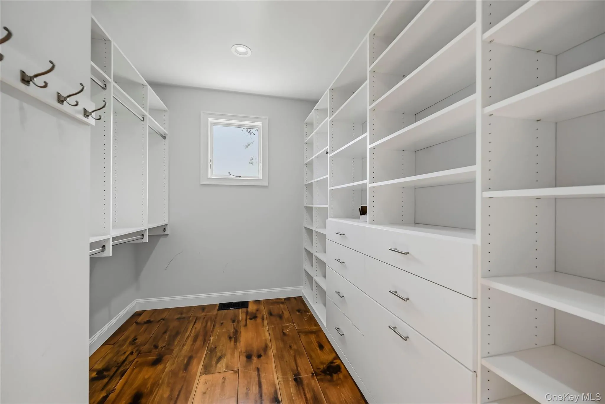 Custom storage organization featuring white floor-to-ceiling shelving, built-in dresser drawers with metallic hardware, integrated garment rods, wall-mounted utility hooks, and wood-finish flooring Custom storage organization featuring white floor-to-ceiling shelving, built-in dresser drawers with metallic hardware, integrated garment rods, wall-mounted utility hooks, and wood-finish flooring