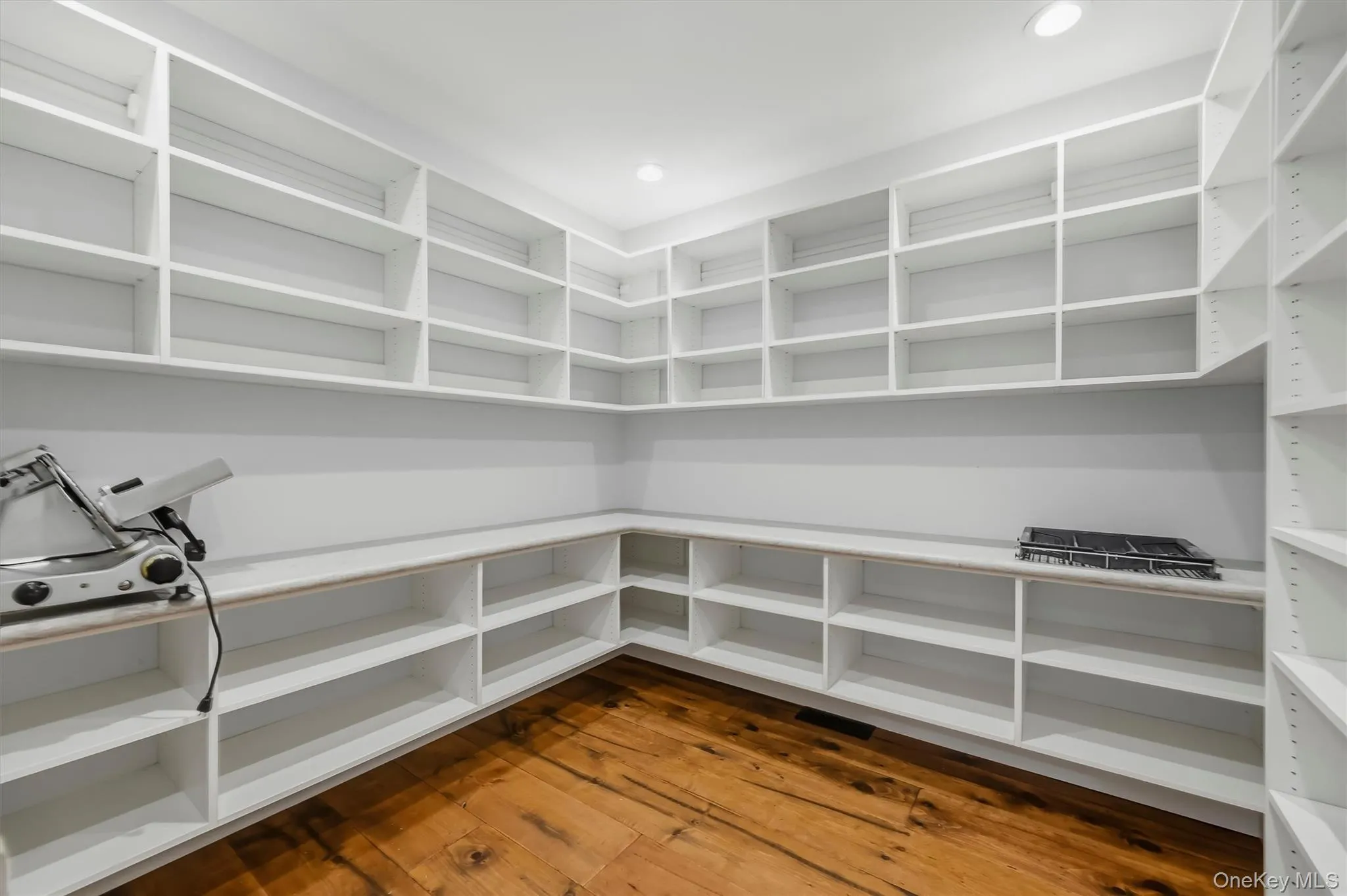 Walk-in pantry featuring floor-to-ceiling white built-in shelving, expansive solid surface countertops, and wide-plank wood-finish flooring Walk-in pantry featuring floor-to-ceiling white built-in shelving, expansive solid surface countertops, and wide-plank wood-finish flooring