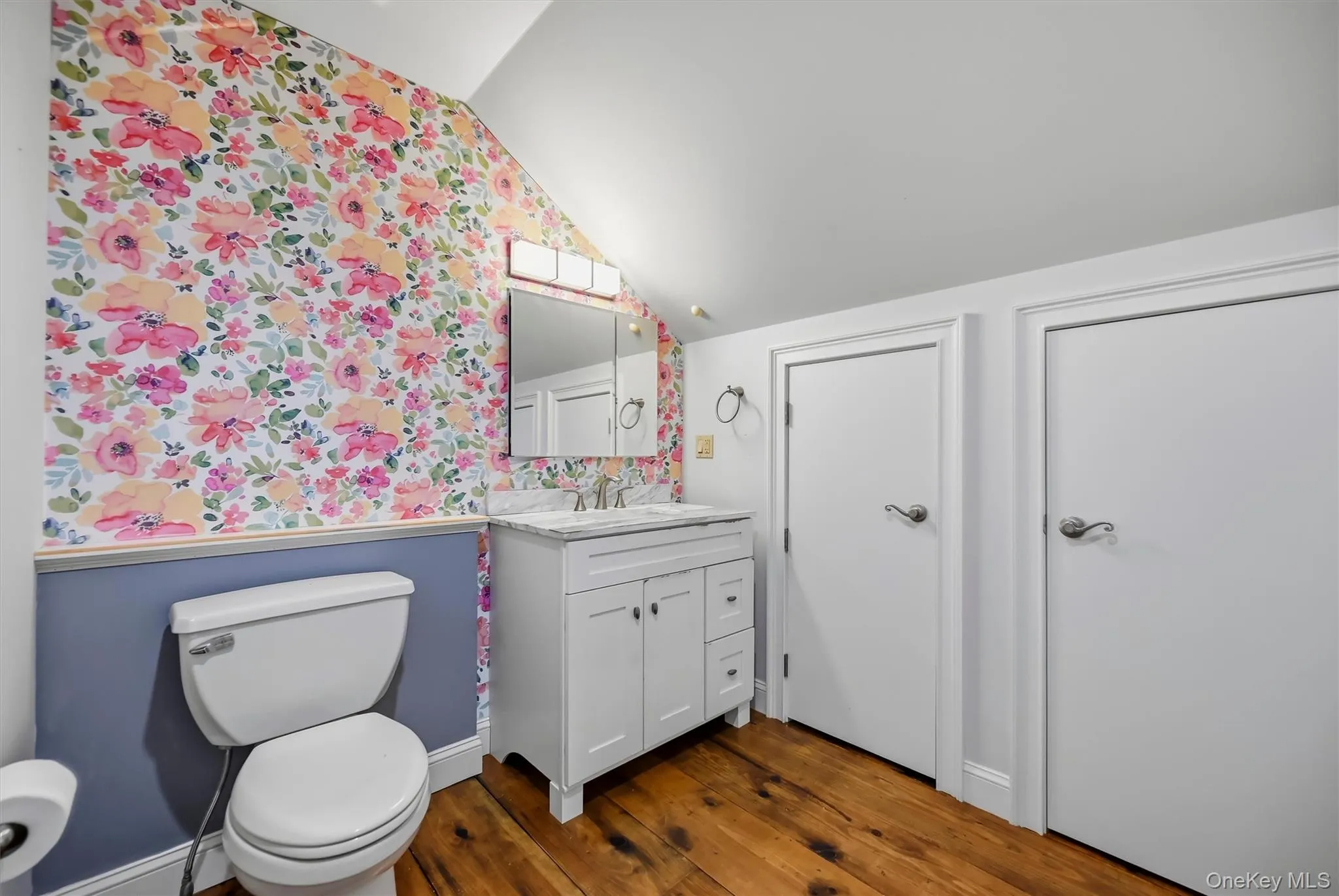 Bathroom featuring wood-finish flooring, floral accent wallpaper, wainscoting, a vanity with a light-toned stone countertop, and a built-in medicine cabinet mirror Bathroom featuring wood-finish flooring, floral accent wallpaper, wainscoting, a vanity with a light-toned stone countertop, and a built-in medicine cabinet mirror