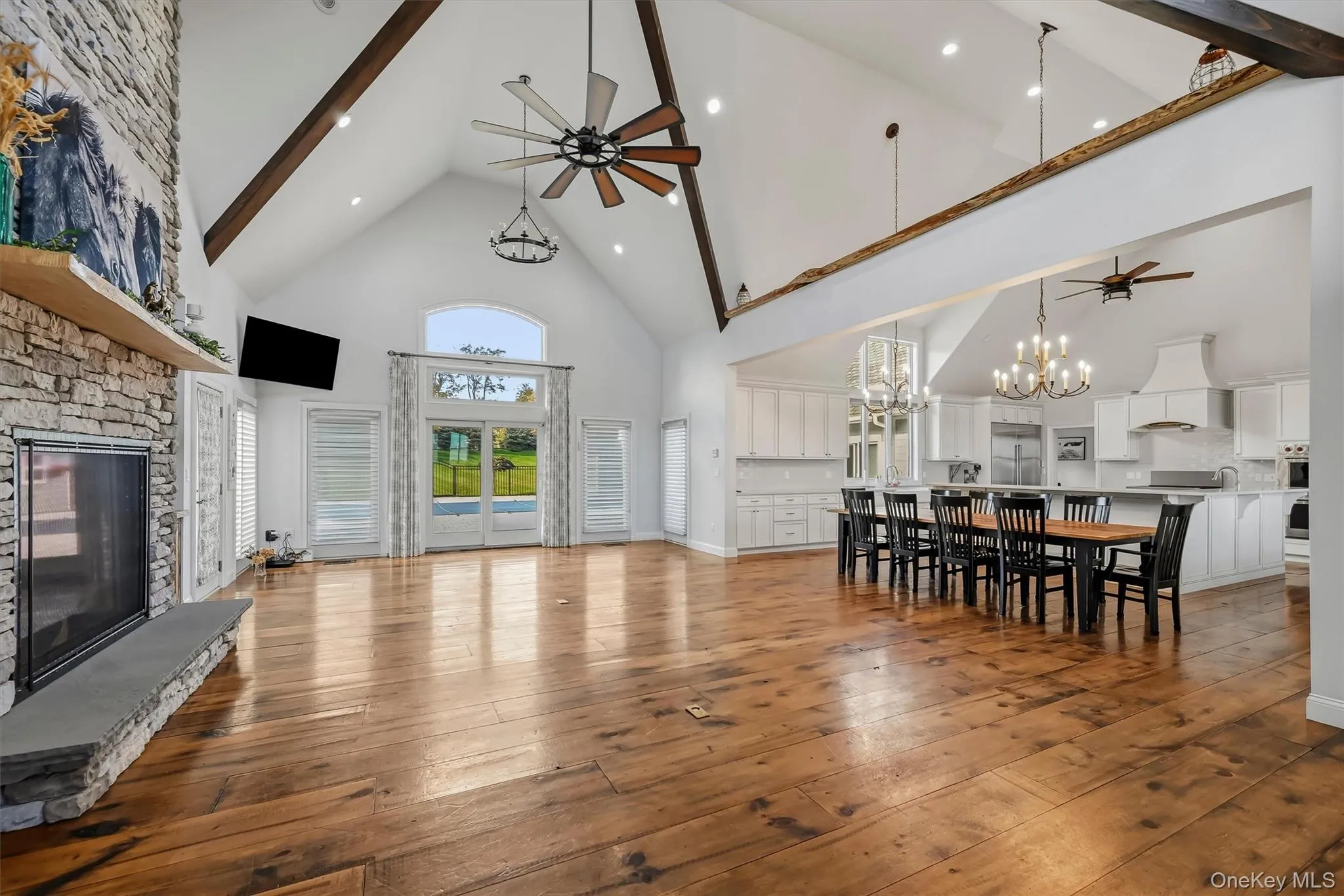 Vaulted ceilings with exposed timber beams, floor-to-ceiling stone fireplace, wide-plank wood-finish flooring, and natural light via arched glass doors Vaulted ceilings with exposed timber beams, floor-to-ceiling stone fireplace, wide-plank wood-finish flooring, and natural light via arched glass doors