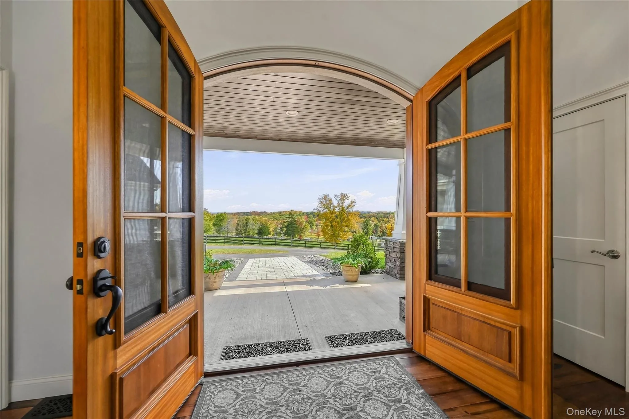 Arched double-entry natural wood doors with glass panels and matte black hardware, opening to a covered porch with a tongue-and-groove ceiling and paver flooring Arched double-entry natural wood doors with glass panels and matte black hardware, opening to a covered porch with a tongue-and-groove ceiling and paver flooring