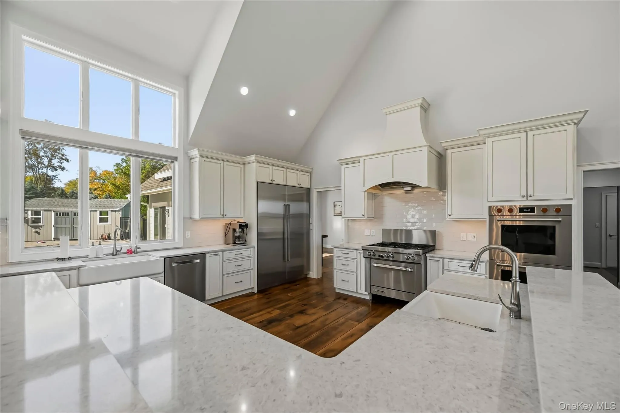 Kitchen featuring high vaulted ceilings, expansive windows, custom white cabinetry, quartz surface countertops, and wood-finish flooring Kitchen featuring high vaulted ceilings, expansive windows, custom white cabinetry, quartz surface countertops, and wood-finish flooring