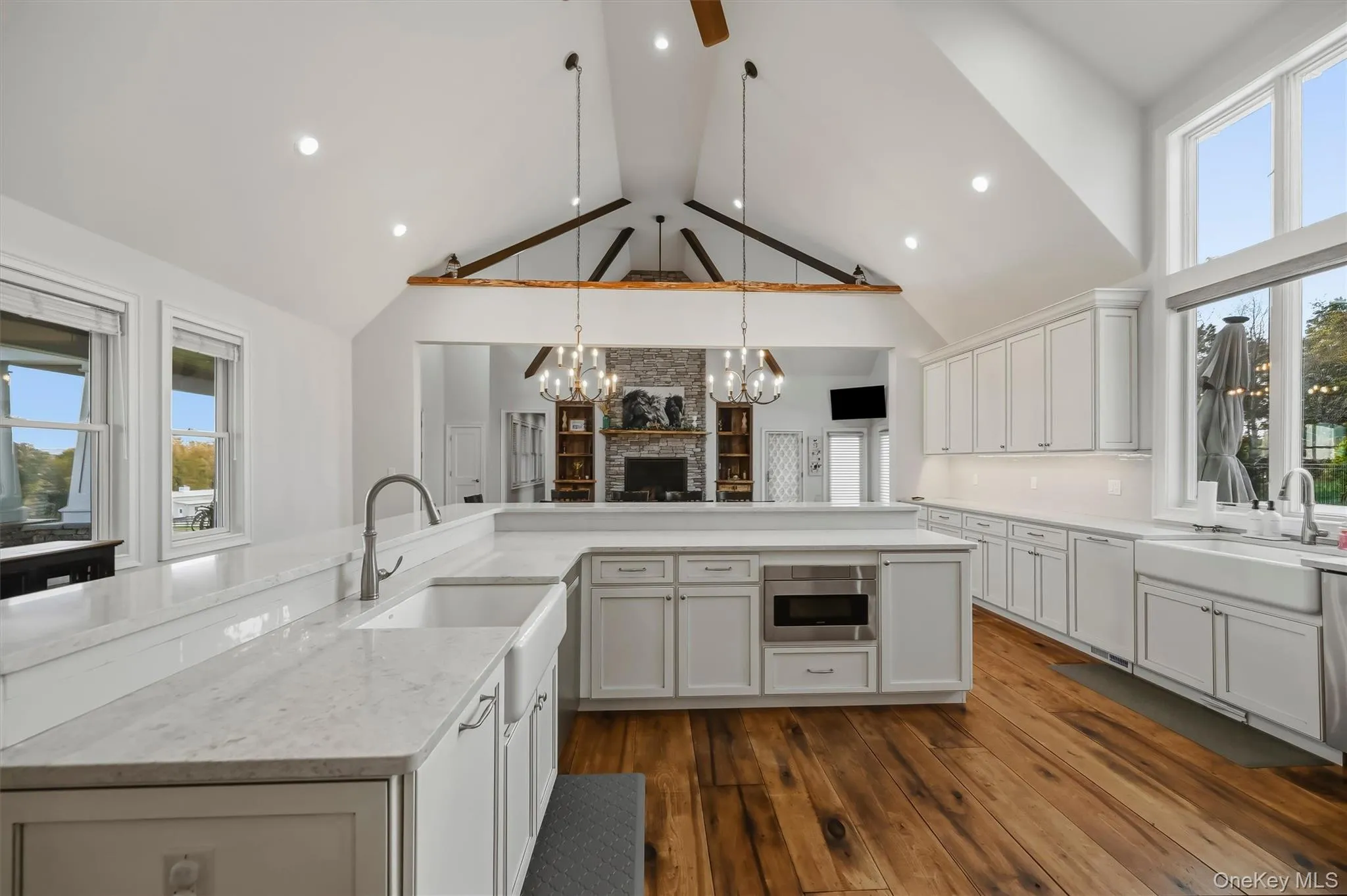 Vaulted ceiling with exposed timber beams, white shaker cabinetry, quartz surfaced countertops, apron-front farmhouse sinks, and wood-finish flooring Vaulted ceiling with exposed timber beams, white shaker cabinetry, quartz surfaced countertops, apron-front farmhouse sinks, and wood-finish flooring
