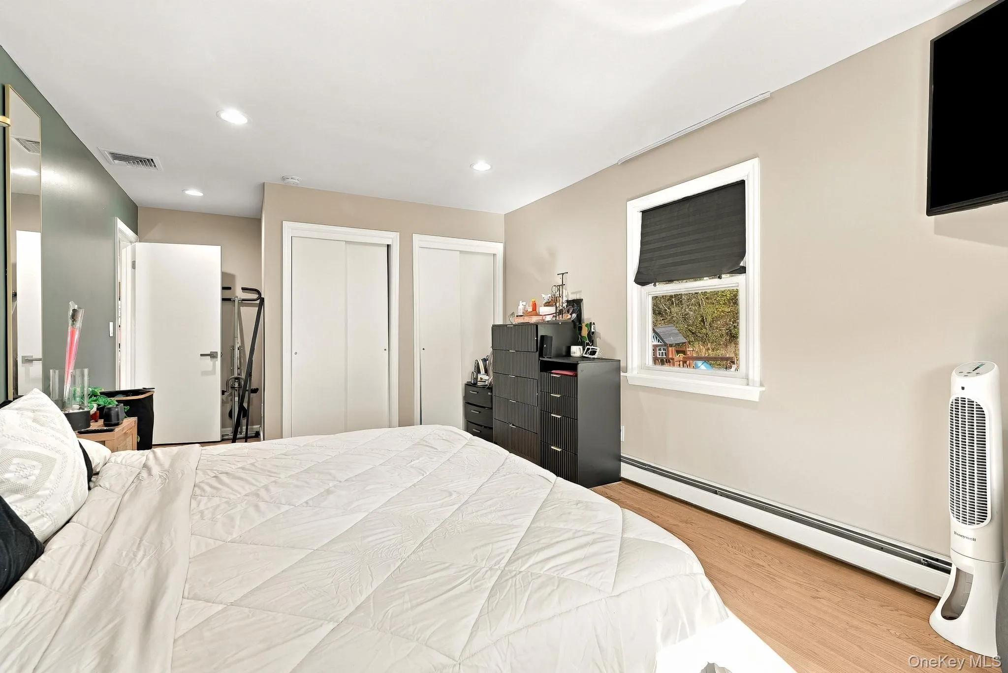 Interior space featuring wood-finish flooring, two sets of bi-fold closet doors, recessed lighting, and a single-hung window with a dark roller shade Interior space featuring wood-finish flooring, two sets of bi-fold closet doors, recessed lighting, and a single-hung window with a dark roller shade
