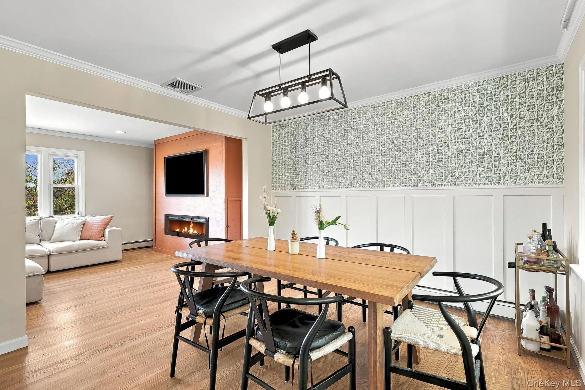 Dining area featuring wood-finish flooring, decorative wall paneling, patterned wallpaper, and a linear four-light metal pendant fixture Dining area featuring wood-finish flooring, decorative wall paneling, patterned wallpaper, and a linear four-light metal pendant fixture