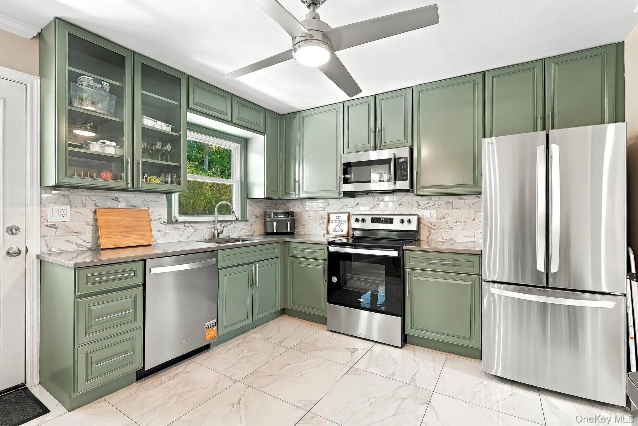 Kitchen featuring sage green cabinetry, stainless steel appliances, stone-patterned tile flooring, light-toned stone-patterned backsplash, and glass-front upper cabinet doors Kitchen featuring sage green cabinetry, stainless steel appliances, stone-patterned tile flooring, light-toned stone-patterned backsplash, and glass-front upper cabinet doors