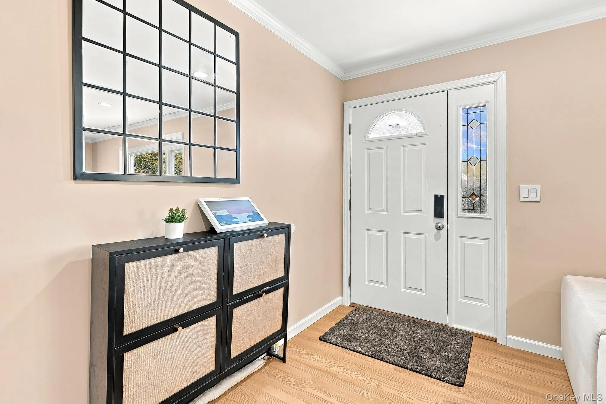 Entryway featuring a white door with a semi-circular transom window and a leaded glass side panel, square grid-pattern wall mirror, and wood-finish flooring Entryway featuring a white door with a semi-circular transom window and a leaded glass side panel, square grid-pattern wall mirror, and wood-finish flooring