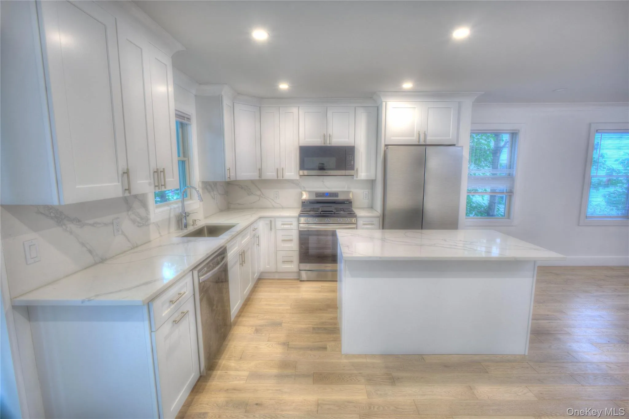 Kitchen featuring white shaker-style cabinetry, stone-finish countertops with integrated backsplash, a central island, stainless steel appliances, and wood-finish flooring Kitchen featuring white shaker-style cabinetry, stone-finish countertops with integrated backsplash, a central island, stainless steel appliances, and wood-finish flooring