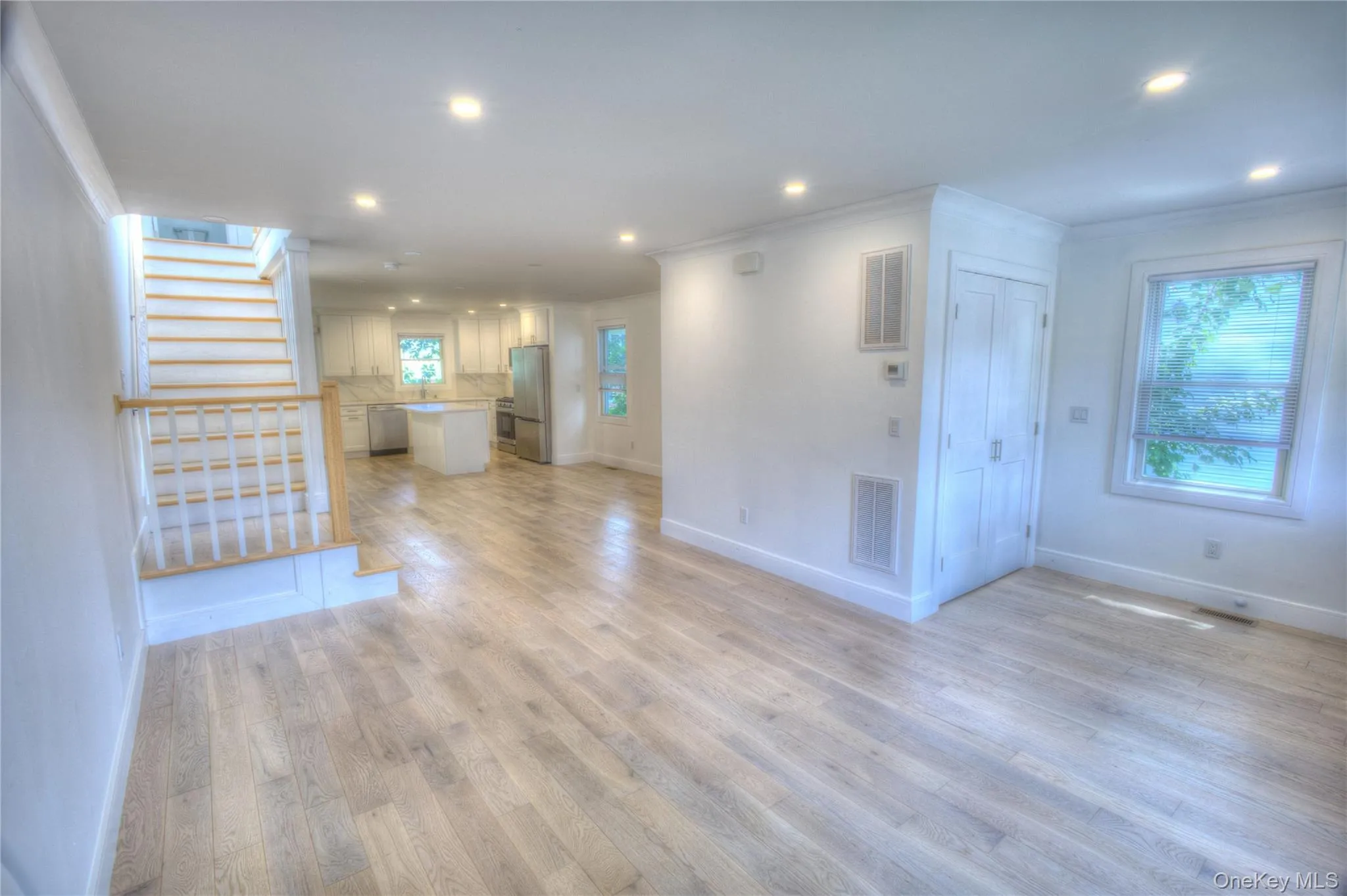 Open-concept living space featuring wood-finish flooring, recessed lighting, a staircase with white balusters and a wood handrail, and white kitchen cabinetry with a central island Open-concept living space featuring wood-finish flooring, recessed lighting, a staircase with white balusters and a wood handrail, and white kitchen cabinetry with a central island