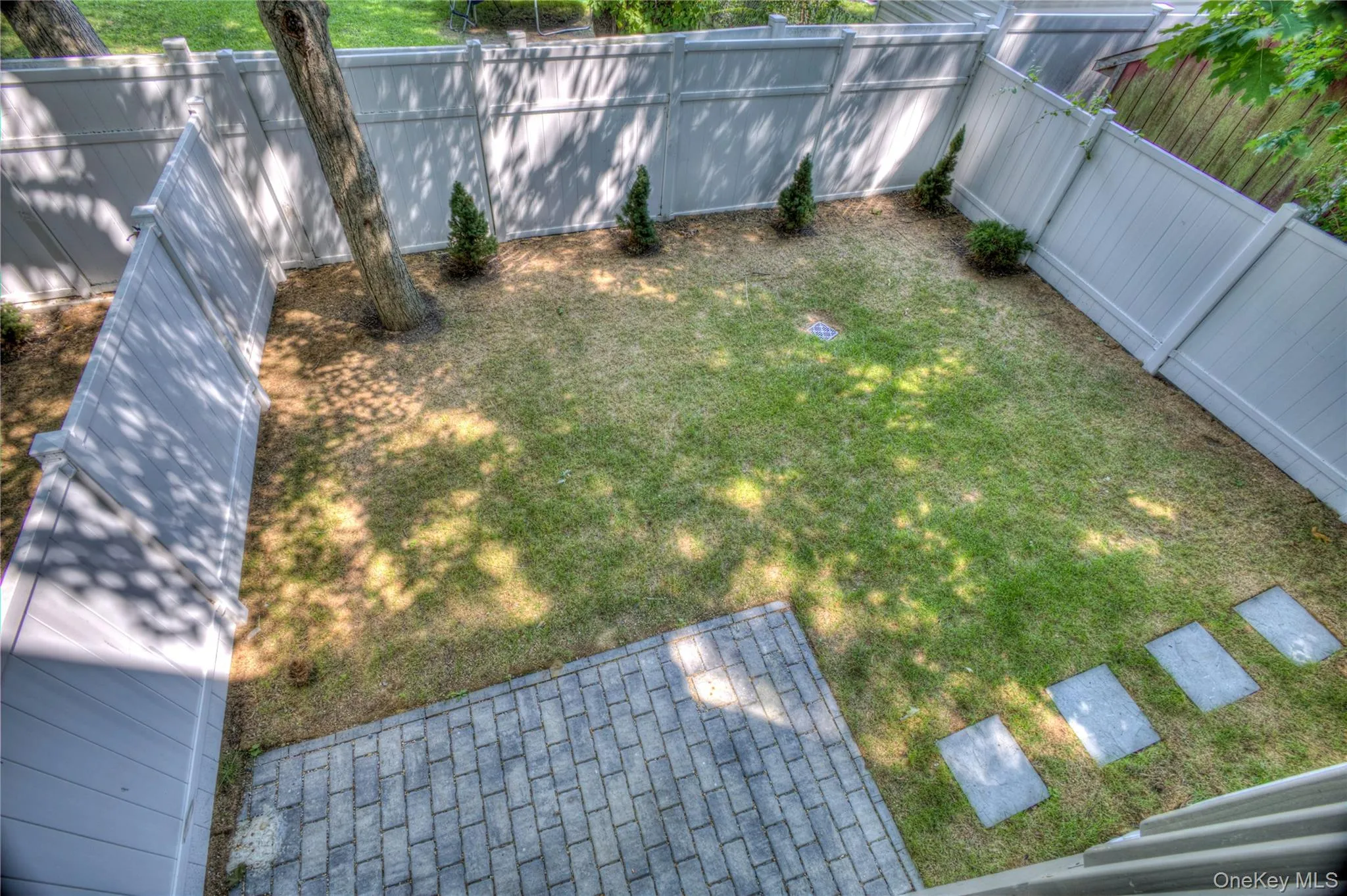 Rear yard featuring a full-height vinyl privacy fence, a grey stone paver patio, a manicured lawn, stone stepping stones, and mature trees Rear yard featuring a full-height vinyl privacy fence, a grey stone paver patio, a manicured lawn, stone stepping stones, and mature trees