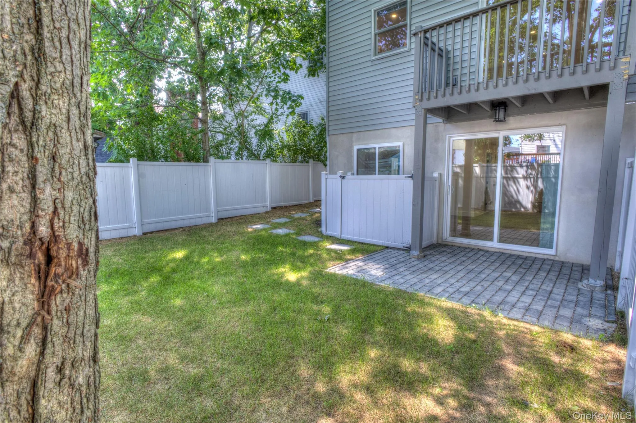 Rear exterior featuring a paver patio, elevated wood balcony, white vinyl privacy fencing, and sliding glass doors Rear exterior featuring a paver patio, elevated wood balcony, white vinyl privacy fencing, and sliding glass doors