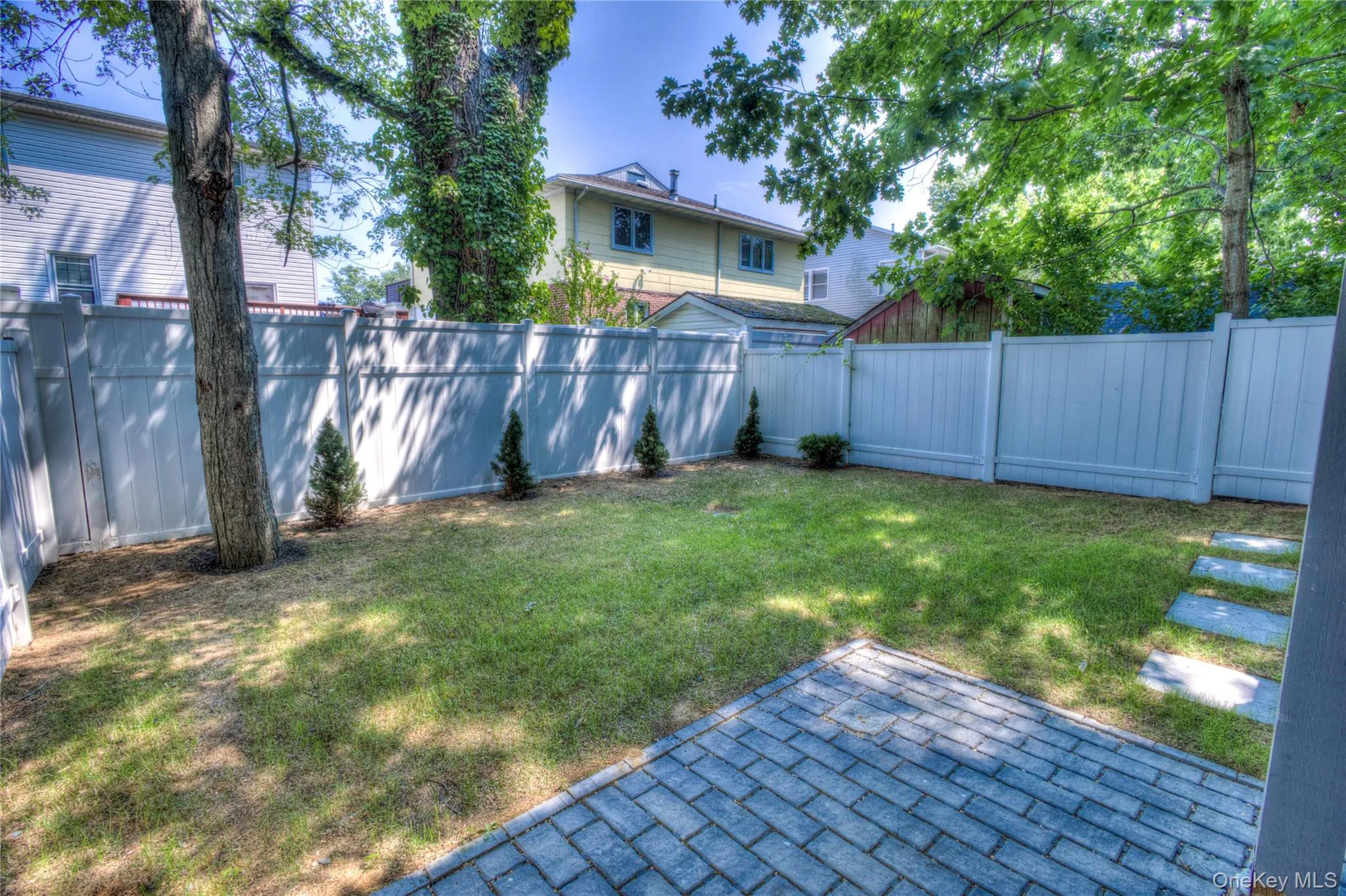 Enclosed backyard featuring a paver patio, stone-finish walkway, white privacy fencing, and mature trees Enclosed backyard featuring a paver patio, stone-finish walkway, white privacy fencing, and mature trees