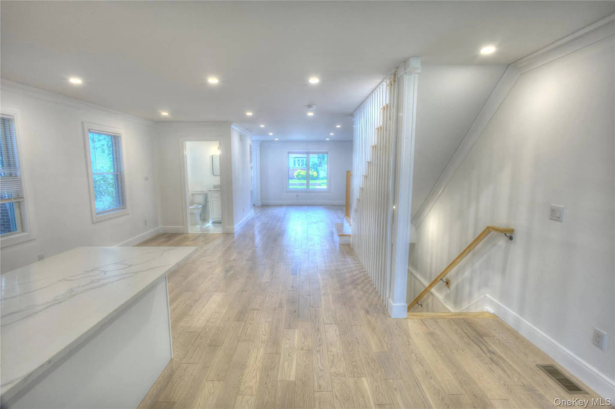 Open-concept interior featuring wood-finish flooring, recessed lighting, a stone-topped kitchen island, and a wooden handrail with vertical balusters Open-concept interior featuring wood-finish flooring, recessed lighting, a stone-topped kitchen island, and a wooden handrail with vertical balusters