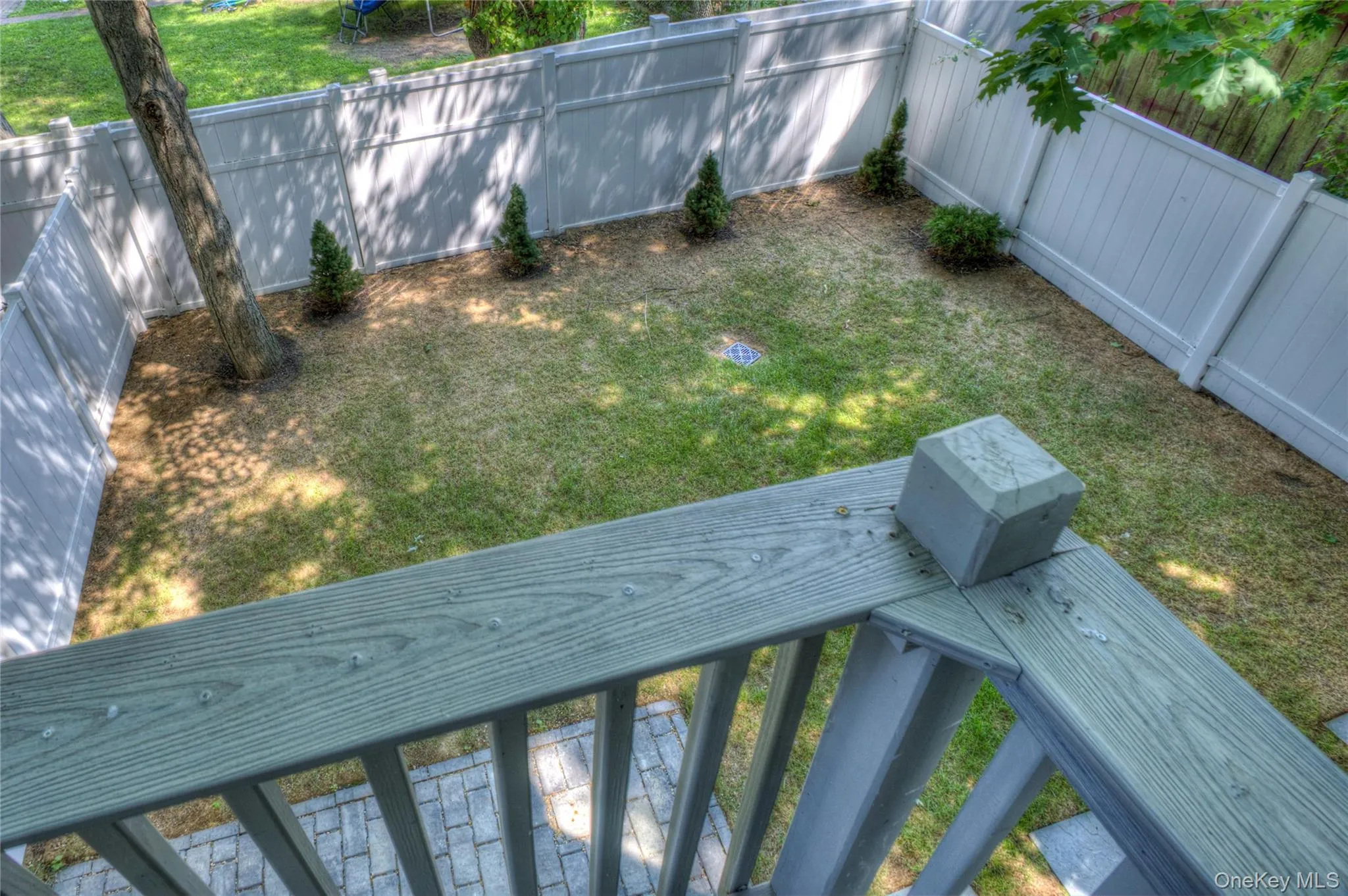 Fenced backyard featuring a wood-tone balcony railing, stone-finish paver patio, lawn area, and mature tree Fenced backyard featuring a wood-tone balcony railing, stone-finish paver patio, lawn area, and mature tree