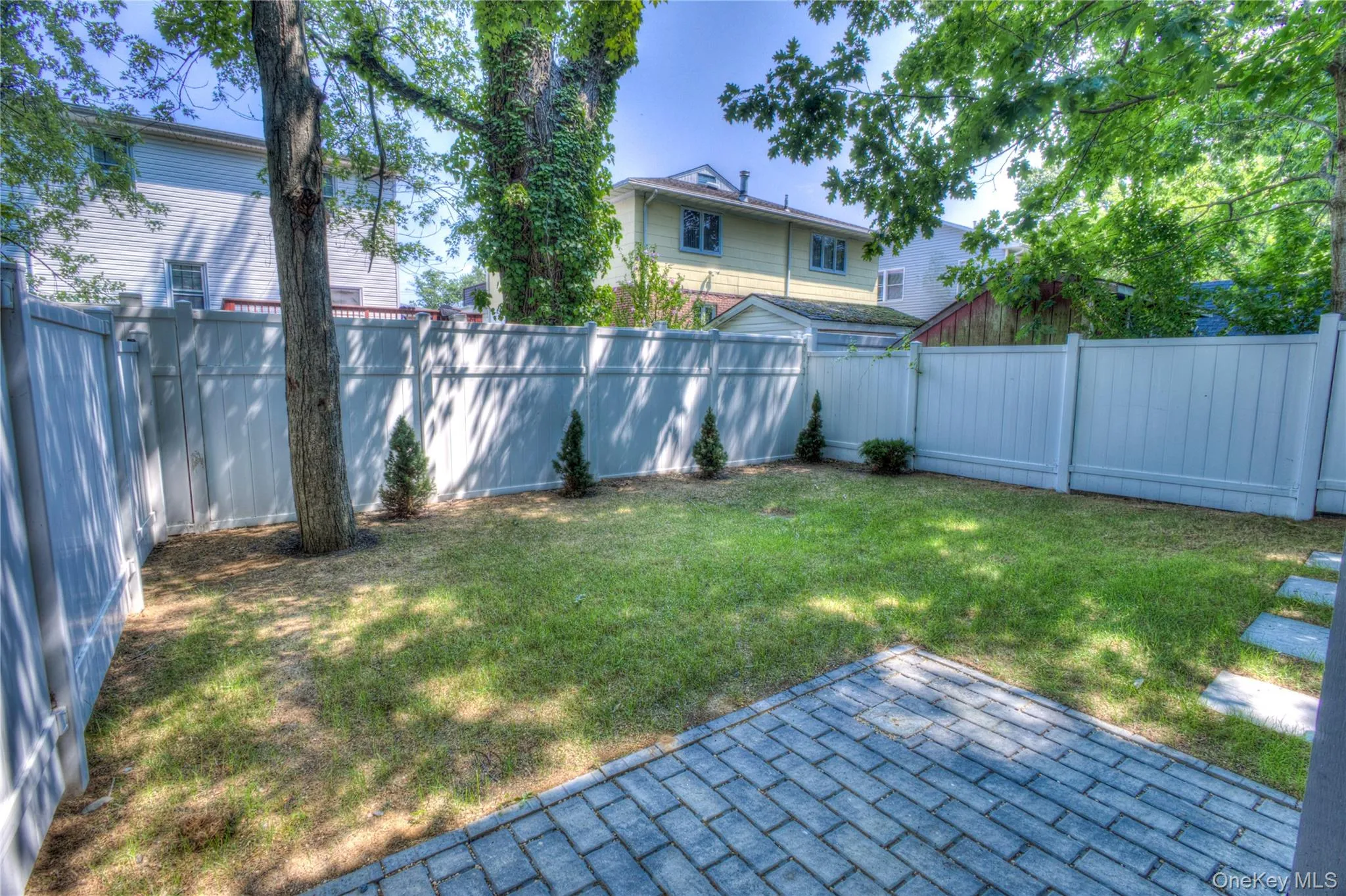 Private backyard featuring a paved stone patio, mature trees, manicured lawn, and full-perimeter vinyl fencing Private backyard featuring a paved stone patio, mature trees, manicured lawn, and full-perimeter vinyl fencing