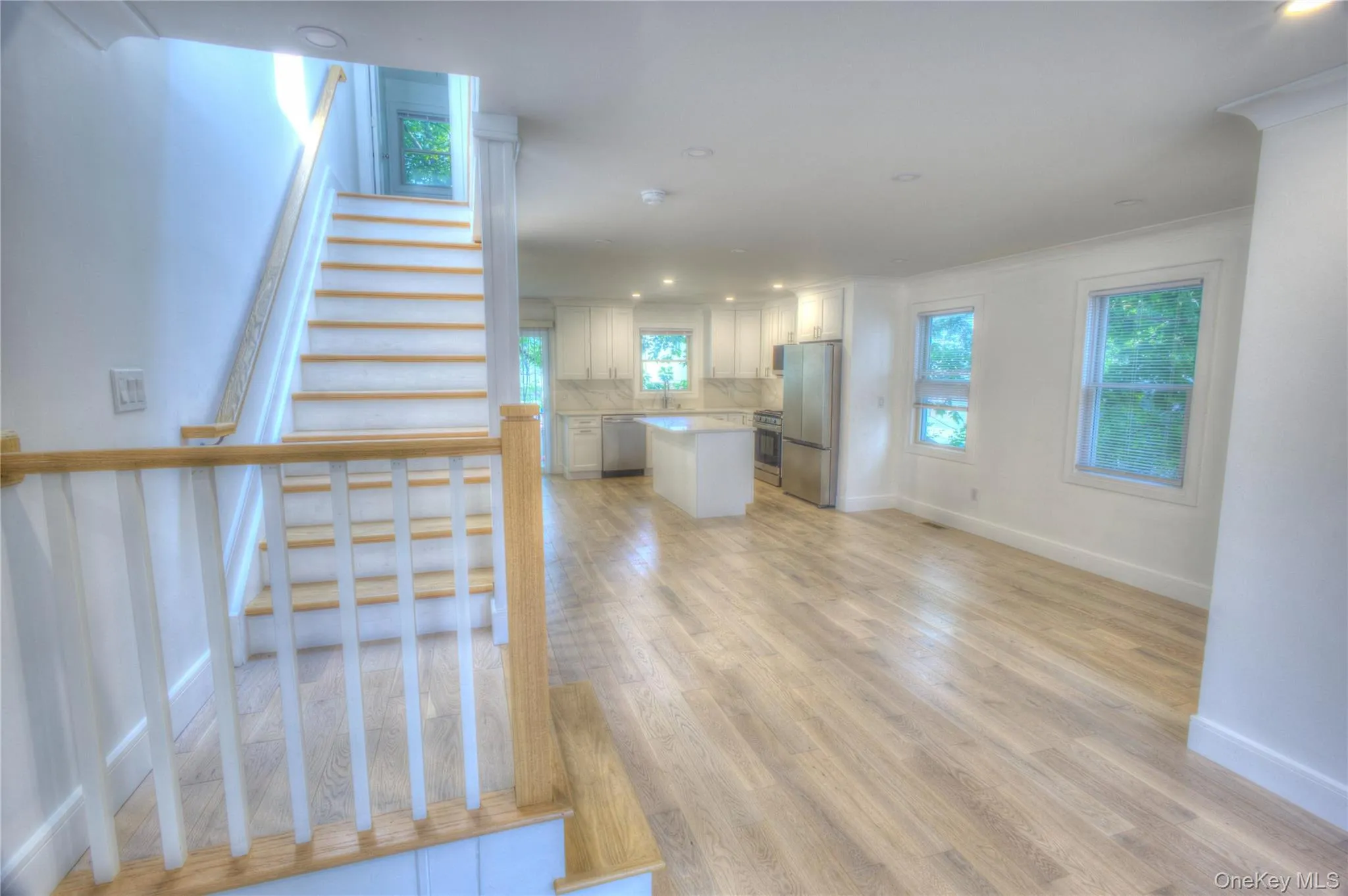 Open-concept interior featuring wood-finish flooring, recessed lighting, and a kitchen equipped with a center island and stainless steel appliances, accented by a staircase with wood treads and a white spindle railing Open-concept interior featuring wood-finish flooring, recessed lighting, and a kitchen equipped with a center island and stainless steel appliances, accented by a staircase with wood treads and a white spindle railing