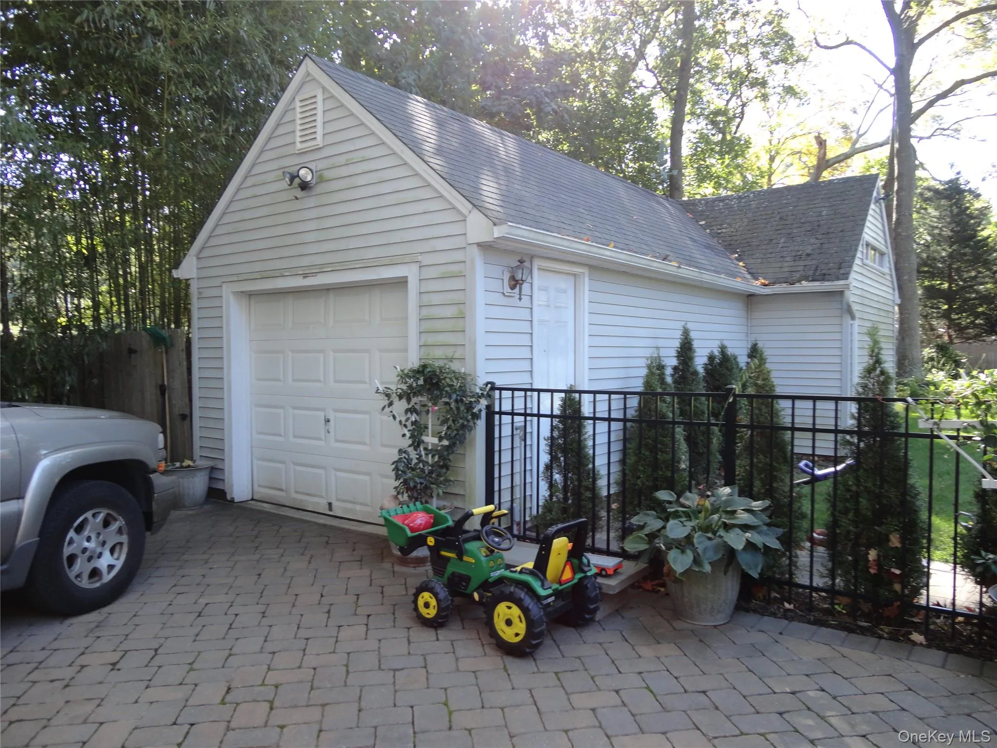 Detached 1.5 garage featuring light-colored horizontal siding, a gabled roof, an overhead garage door, and decorative wall-mounted lighting, set on a herringbone-patterned paver driveway with black metal perimeter fencing Detached 1.5 garage featuring light-colored horizontal siding, a gabled roof, an overhead garage door, and decorative wall-mounted lighting, set on a herringbone-patterned paver driveway with black metal perimeter fencing