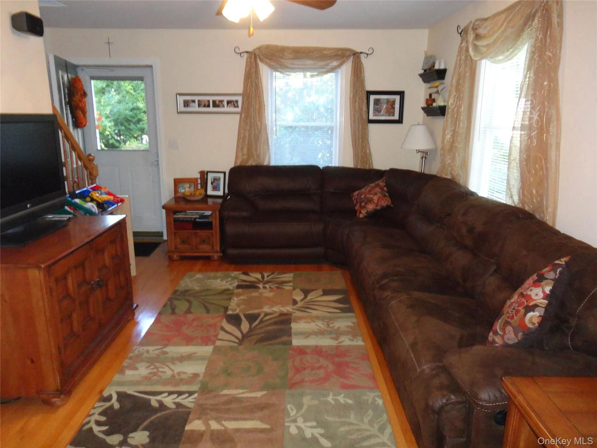 Living area featuring wood-finish flooring, two large windows with decorative drapery hardware, a ceiling fan, and an exterior door Living area featuring wood-finish flooring, two large windows with decorative drapery hardware, a ceiling fan, and an exterior door