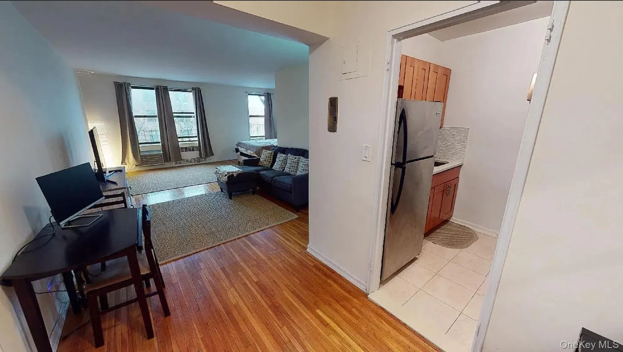 Open-plan living area featuring wood-finish flooring and multiple windows, paired with a separate kitchen containing stainless steel appliances and light-colored tile flooring Open-plan living area featuring wood-finish flooring and multiple windows, paired with a separate kitchen containing stainless steel appliances and light-colored tile flooring