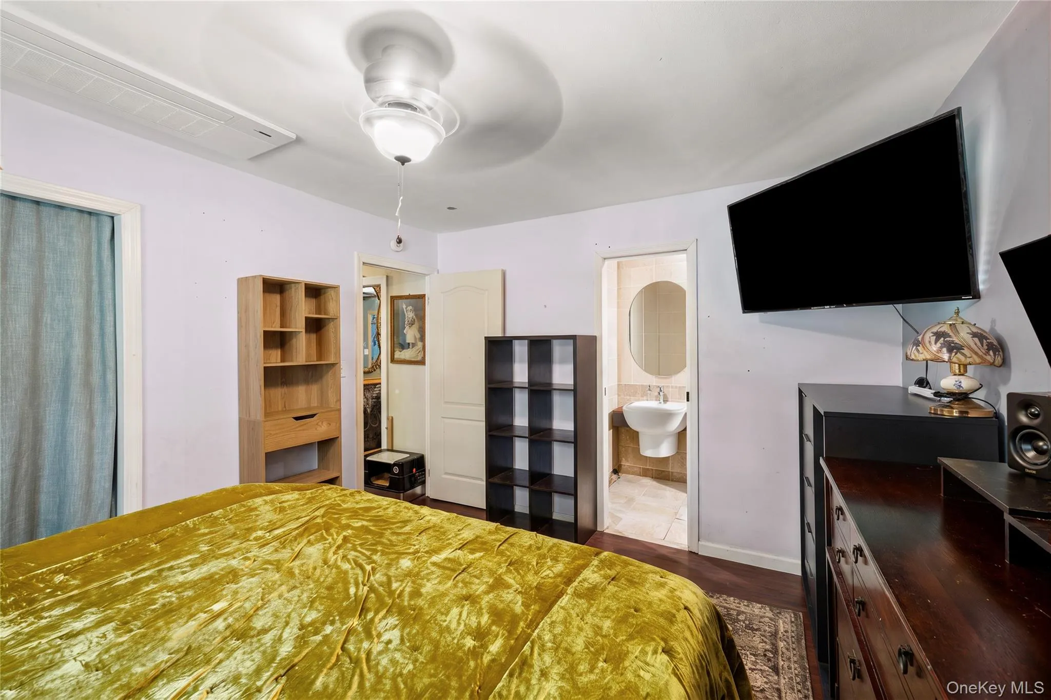 Room featuring dark wood-finish flooring, a ceiling fan with integrated lighting, and a wall-mounted interior door leading to a bathroom with a pedestal sink and circular mirror (Decluttered Edit) Room featuring dark wood-finish flooring, a ceiling fan with integrated lighting, and a wall-mounted interior door leading to a bathroom with a pedestal sink and circular mirror (Decluttered Edit)