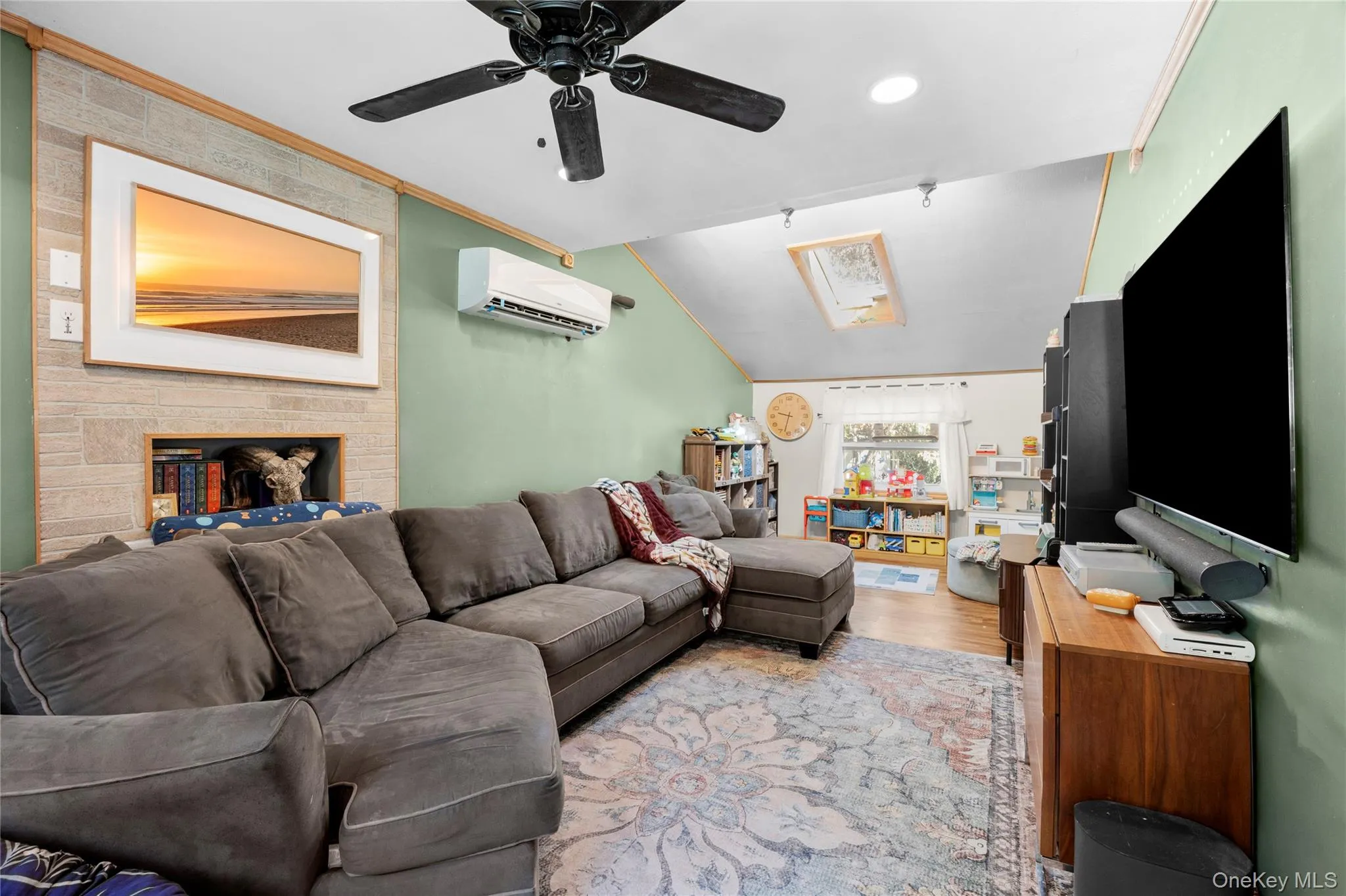 Vaulted ceiling featuring a skylight, stone-faced fireplace, wall-mounted ductless heating and cooling unit, ceiling fan, and wood-finish flooring Vaulted ceiling featuring a skylight, stone-faced fireplace, wall-mounted ductless heating and cooling unit, ceiling fan, and wood-finish flooring