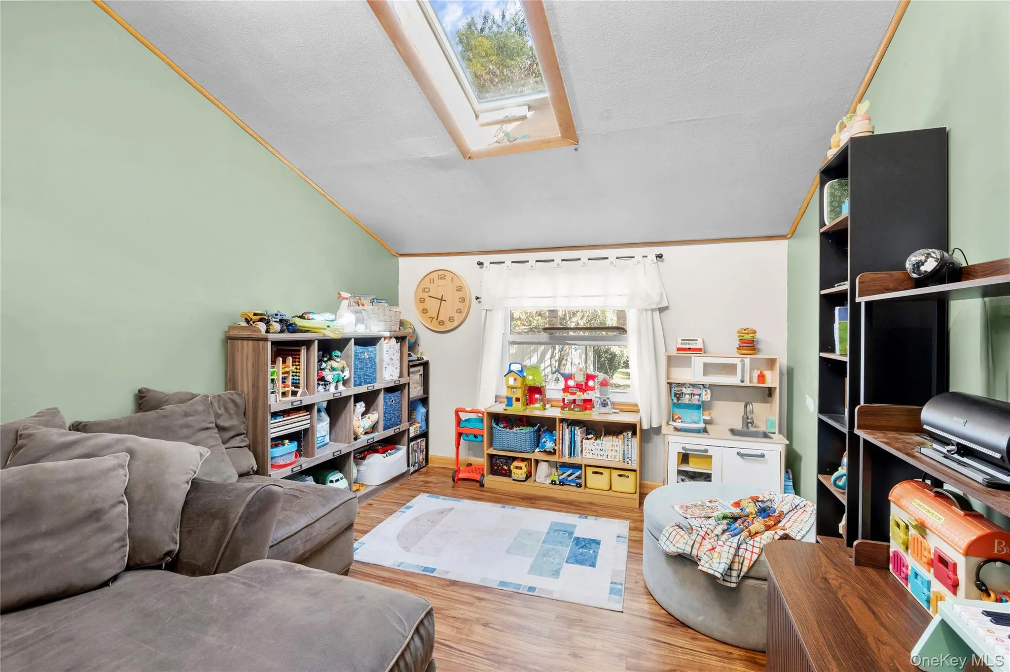 Vaulted ceiling featuring a large rectangular skylight, wood-finish flooring, and natural light from a window with decorative trim Vaulted ceiling featuring a large rectangular skylight, wood-finish flooring, and natural light from a window with decorative trim