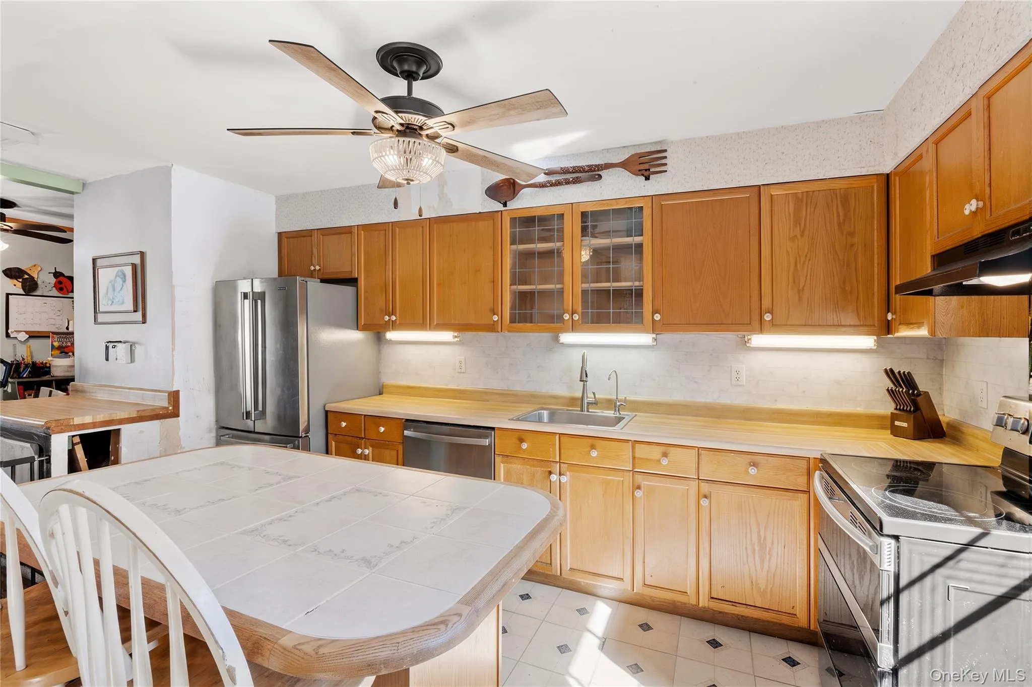 Kitchen featuring wood cabinetry, glass-paneled upper cabinets, tile-topped center island, stainless steel refrigerator, and built-in under-cabinet lighting (Decluttered Edit) Kitchen featuring wood cabinetry, glass-paneled upper cabinets, tile-topped center island, stainless steel refrigerator, and built-in under-cabinet lighting (Decluttered Edit)