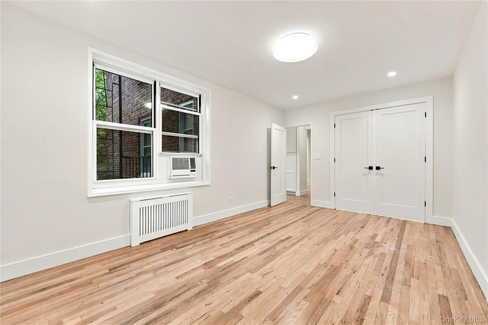Interior space featuring wood-finish flooring, double-panel closet doors with matte black hardware, recessed lighting, and a window with a metal radiator cover Interior space featuring wood-finish flooring, double-panel closet doors with matte black hardware, recessed lighting, and a window with a metal radiator cover