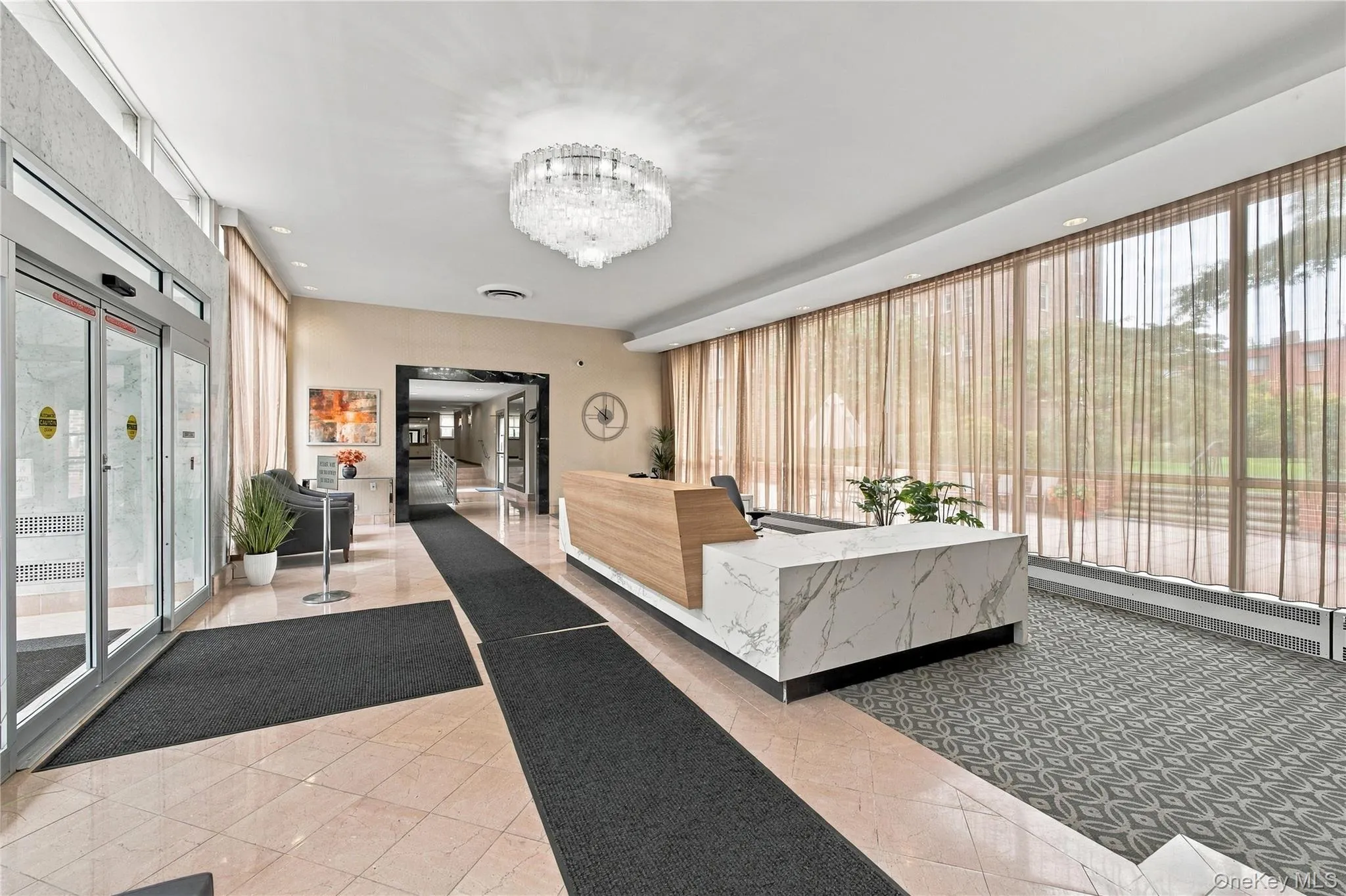 Grand lobby area featuring polished tile flooring, a stone-finish reception desk with wood detailing, floor-to-ceiling windows, and a tiered crystal chandelier Grand lobby area featuring polished tile flooring, a stone-finish reception desk with wood detailing, floor-to-ceiling windows, and a tiered crystal chandelier