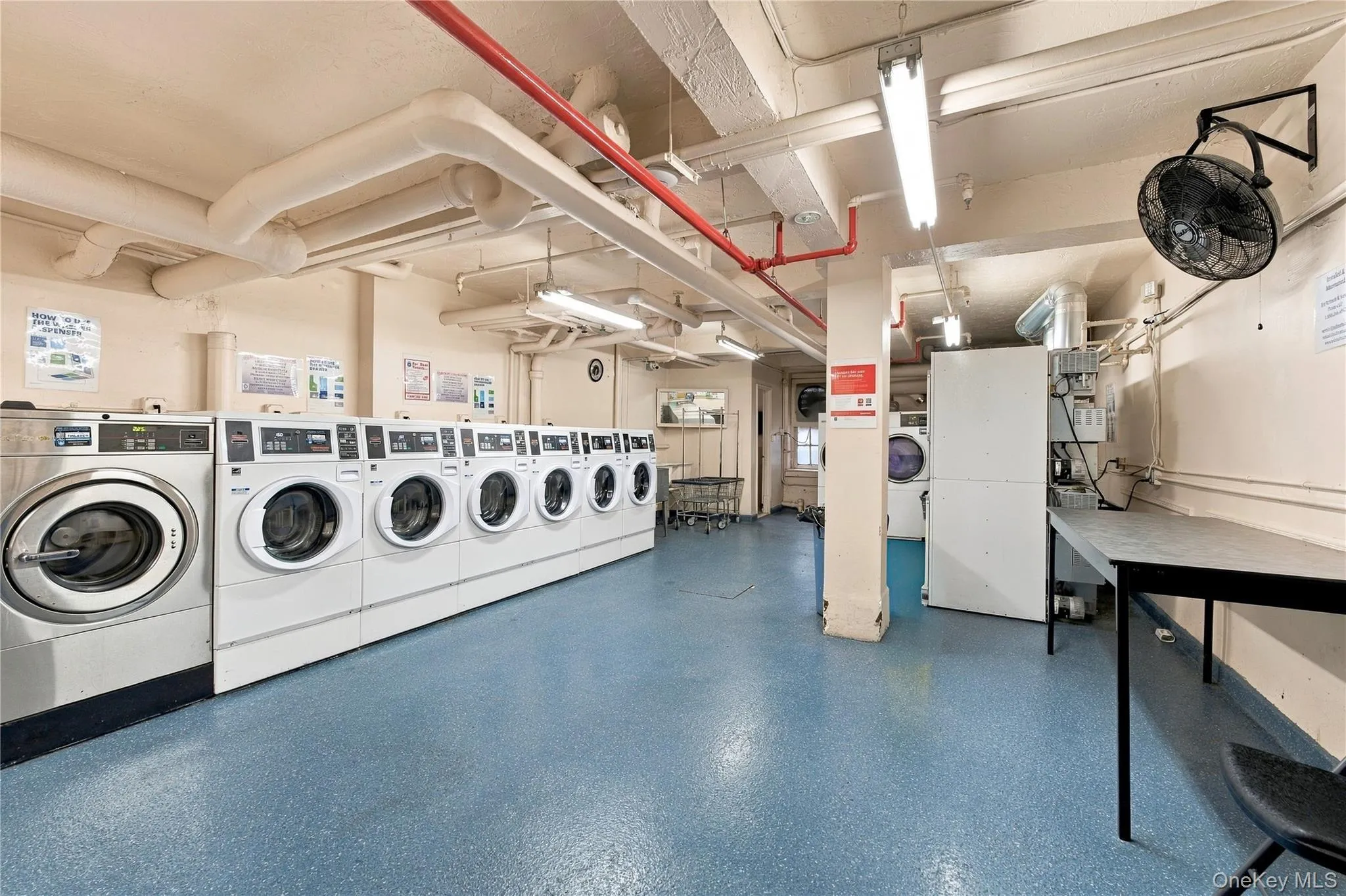 Laundry facility featuring multiple front-loading washers, overhead linear lighting, durable industrial-style flooring, and a built-in folding surface Laundry facility featuring multiple front-loading washers, overhead linear lighting, durable industrial-style flooring, and a built-in folding surface