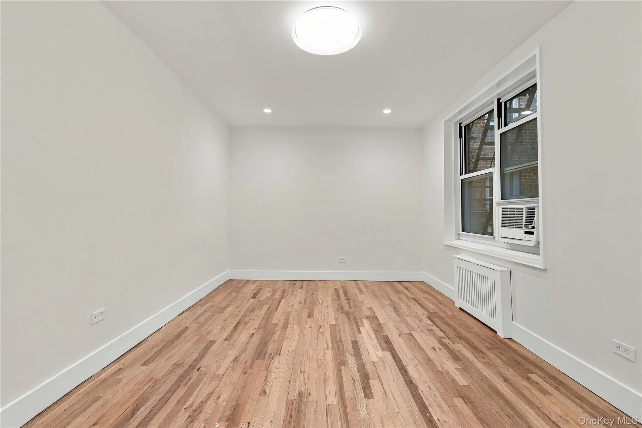 Interior space featuring wood-finish flooring, white walls, recessed ceiling lighting, a flush-mount overhead light fixture, and a window with a built-in radiator enclosure Interior space featuring wood-finish flooring, white walls, recessed ceiling lighting, a flush-mount overhead light fixture, and a window with a built-in radiator enclosure