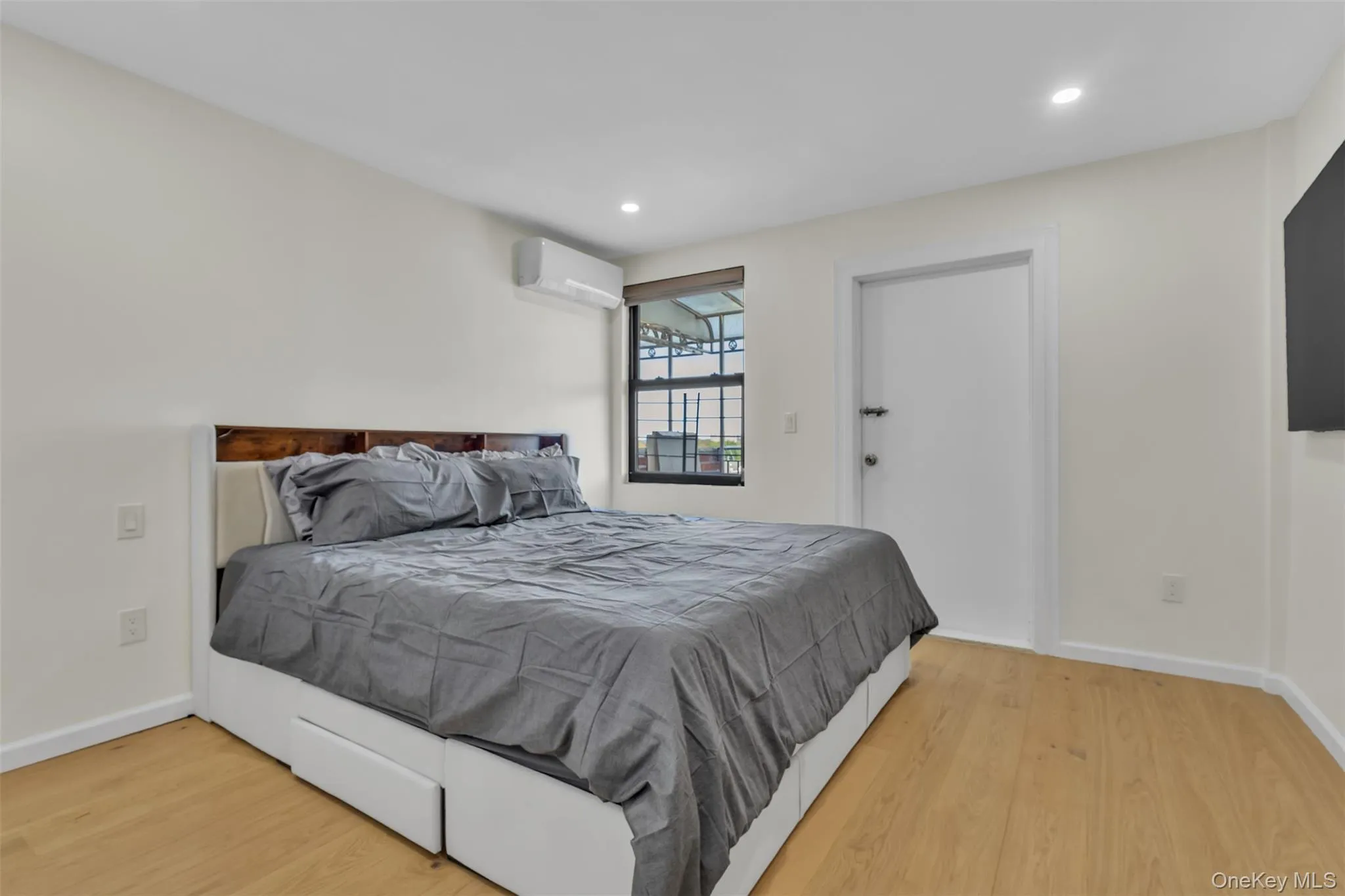 Room featuring neutral wall paint, wood-finish flooring, recessed lighting, a window with integrated shade, and a wall-mounted ductless climate control unit Room featuring neutral wall paint, wood-finish flooring, recessed lighting, a window with integrated shade, and a wall-mounted ductless climate control unit