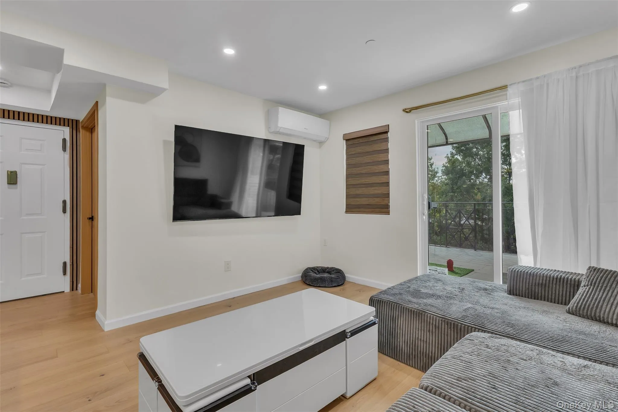 Light-toned wood-finish flooring, recessed lighting, wall-mounted ductless heating and cooling unit, and glass sliding door opening to an exterior balcony Light-toned wood-finish flooring, recessed lighting, wall-mounted ductless heating and cooling unit, and glass sliding door opening to an exterior balcony