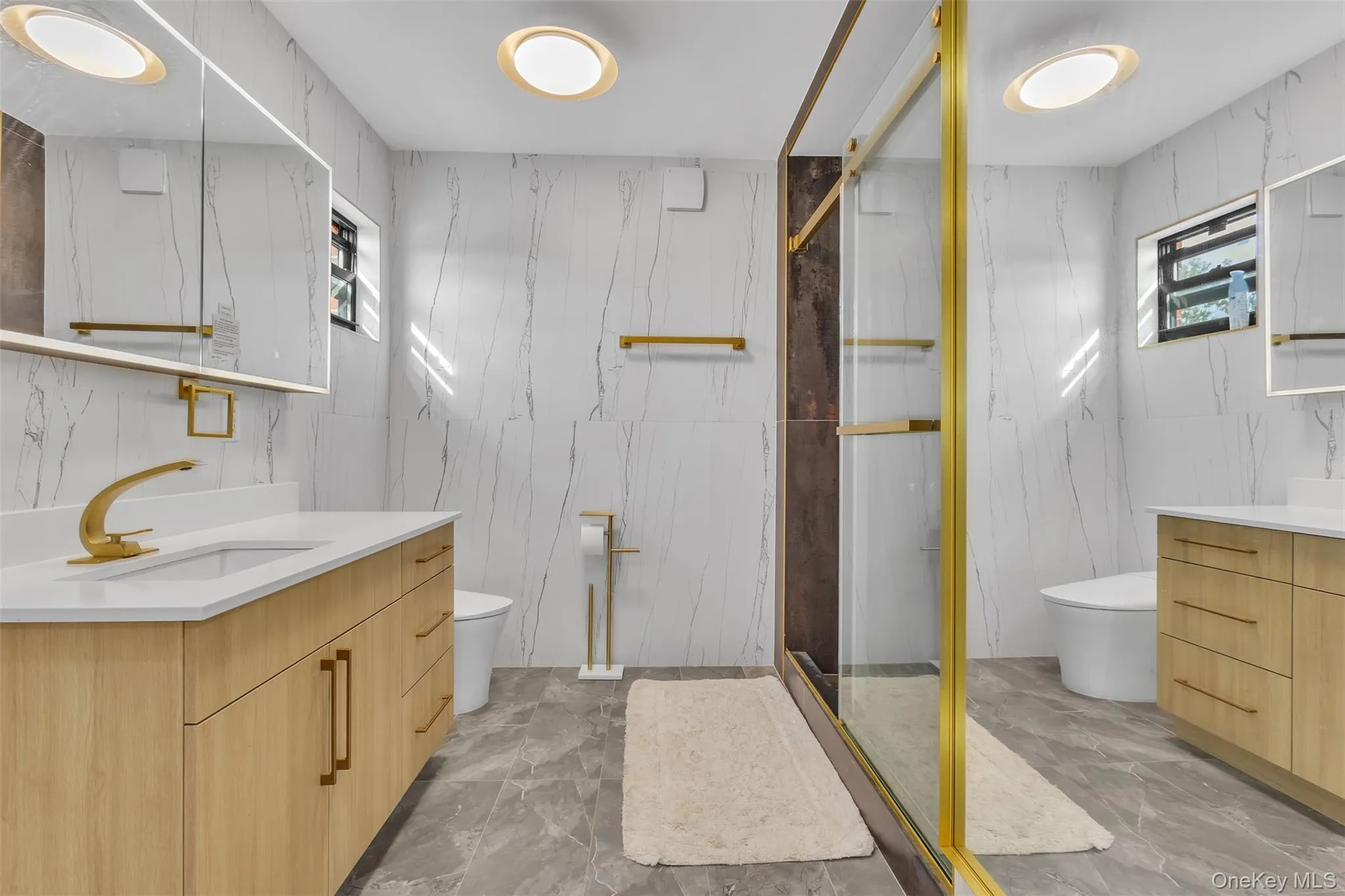 Bathroom featuring light wood-finish vanities with rectangular integrated sinks, gold-tone fixtures, floor-to-ceiling white tiled walls with veining, a glass-enclosed shower with gold-tone framing, and large-format tile flooring Bathroom featuring light wood-finish vanities with rectangular integrated sinks, gold-tone fixtures, floor-to-ceiling white tiled walls with veining, a glass-enclosed shower with gold-tone framing, and large-format tile flooring