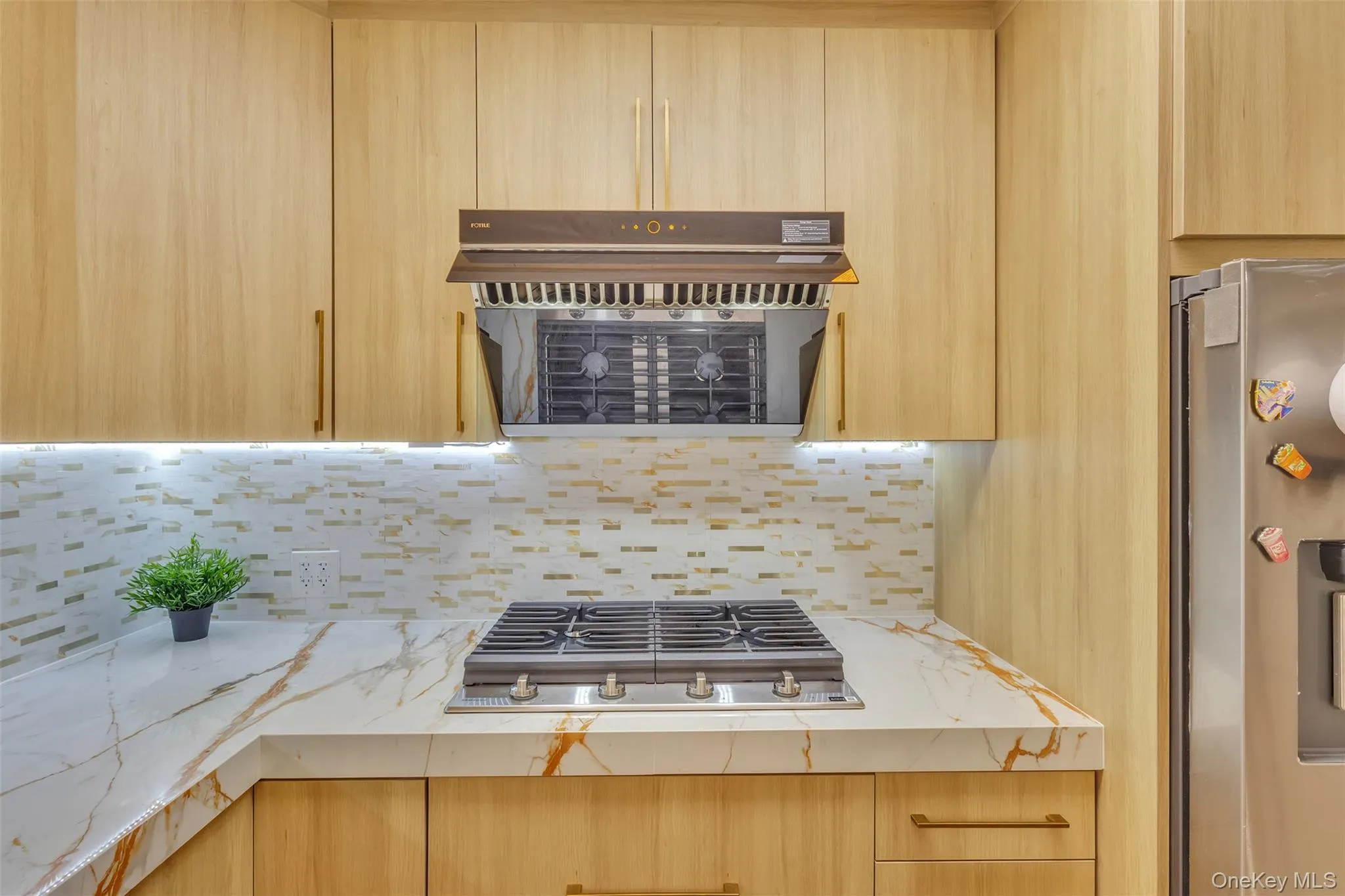 Kitchen featuring light wood-finish cabinetry, stone-patterned countertops, a tile backsplash, a stainless steel gas cooktop, and a matching range hood Kitchen featuring light wood-finish cabinetry, stone-patterned countertops, a tile backsplash, a stainless steel gas cooktop, and a matching range hood