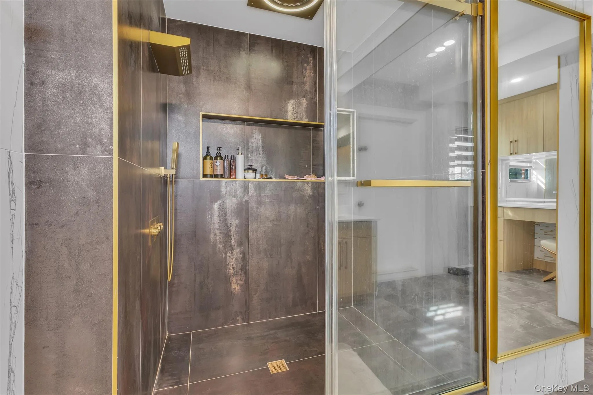 Walk-in shower featuring large-format metallic-finish wall tiles, built-in recessed shelving, gold-tone hardware, and a frameless glass door enclosure Walk-in shower featuring large-format metallic-finish wall tiles, built-in recessed shelving, gold-tone hardware, and a frameless glass door enclosure