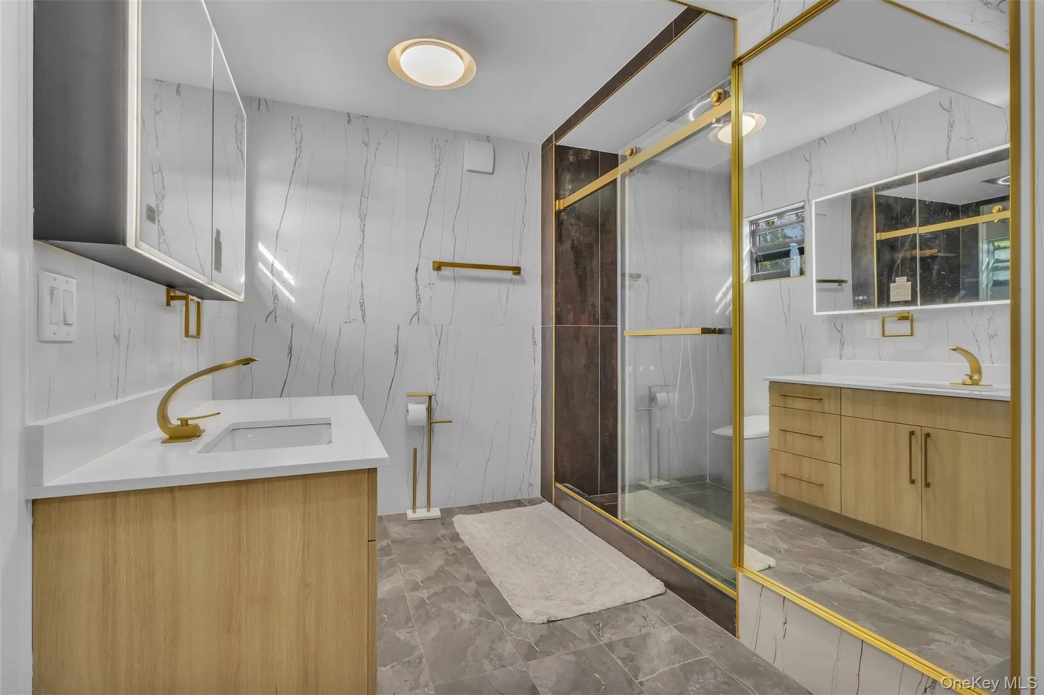 Bathroom featuring wood-finish cabinetry, white solid-surface countertops, gold-toned hardware, full-height tile wall cladding, and a glass-enclosed shower Bathroom featuring wood-finish cabinetry, white solid-surface countertops, gold-toned hardware, full-height tile wall cladding, and a glass-enclosed shower