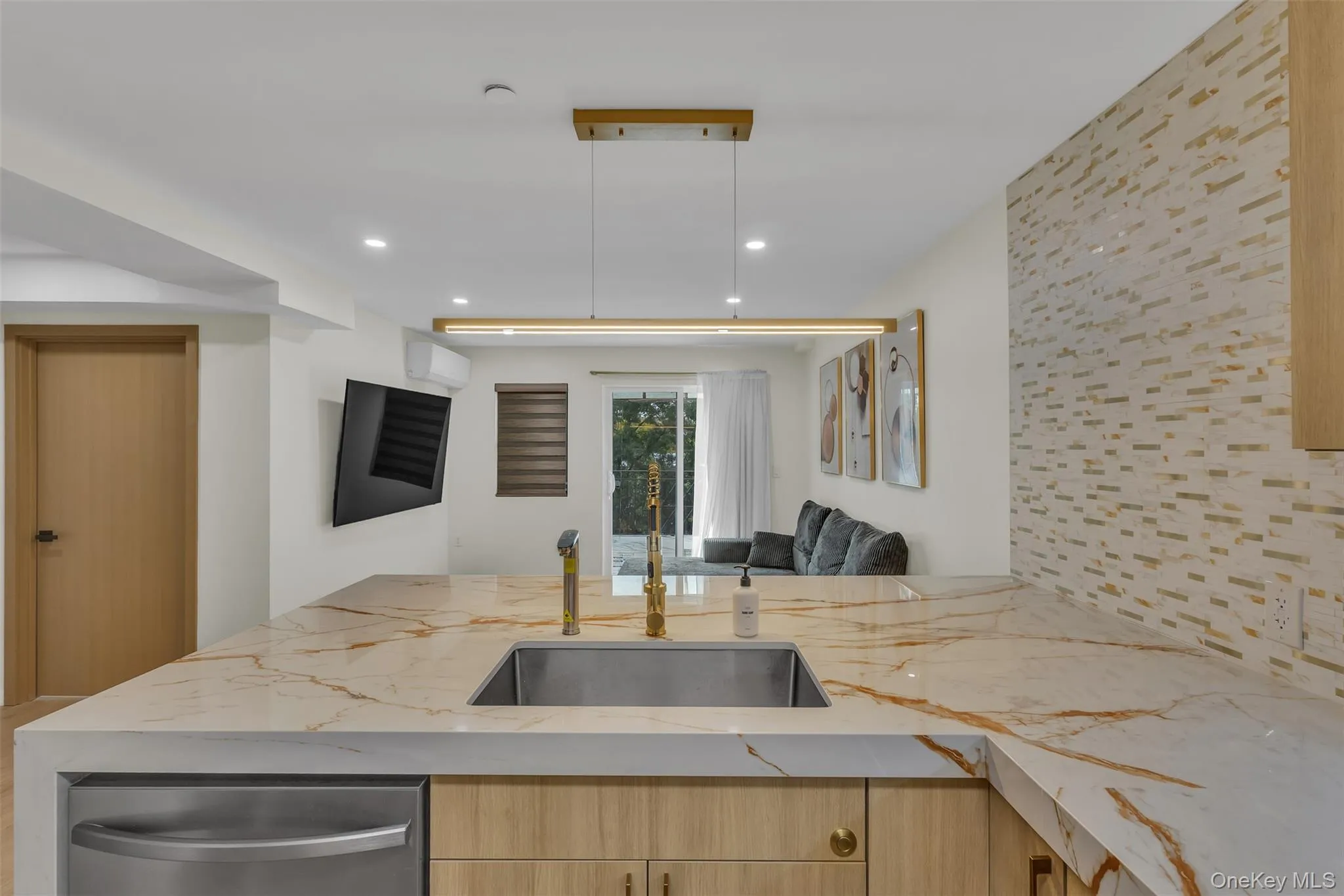 Kitchen island with stone-finish countertop, undermount stainless steel sink, gold-tone faucet, linear pendant lighting, and textured tile backsplash Kitchen island with stone-finish countertop, undermount stainless steel sink, gold-tone faucet, linear pendant lighting, and textured tile backsplash