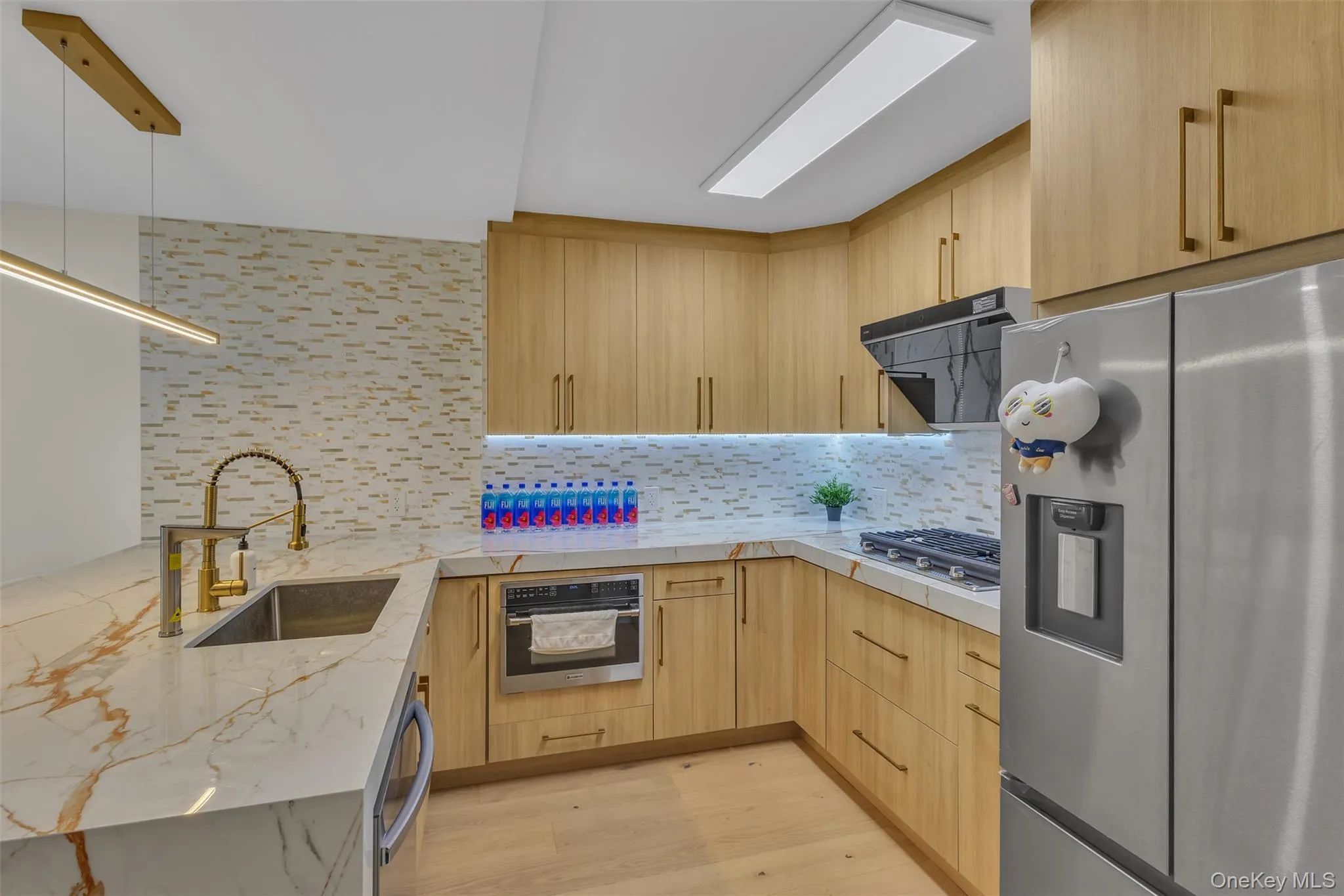 Kitchen featuring light wood-finish cabinetry, stone-patterned countertops, a textured mosaic backsplash, an undermount stainless steel sink with a high-arch gold-tone faucet, and built-in stainless steel appliances Kitchen featuring light wood-finish cabinetry, stone-patterned countertops, a textured mosaic backsplash, an undermount stainless steel sink with a high-arch gold-tone faucet, and built-in stainless steel appliances