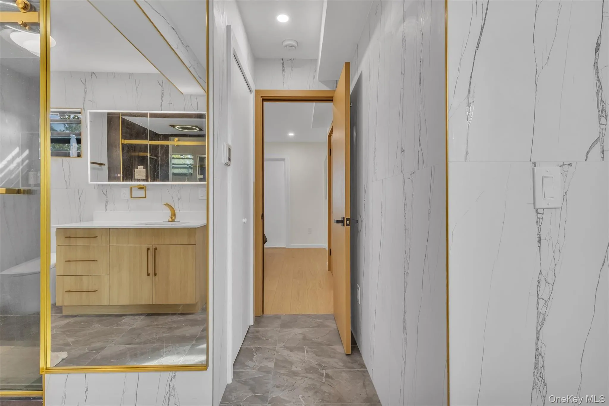 Bathroom featuring large-format wall tiles with veining, wood-finish cabinetry with metallic hardware, recessed lighting, and a gold-trimmed mirror Bathroom featuring large-format wall tiles with veining, wood-finish cabinetry with metallic hardware, recessed lighting, and a gold-trimmed mirror