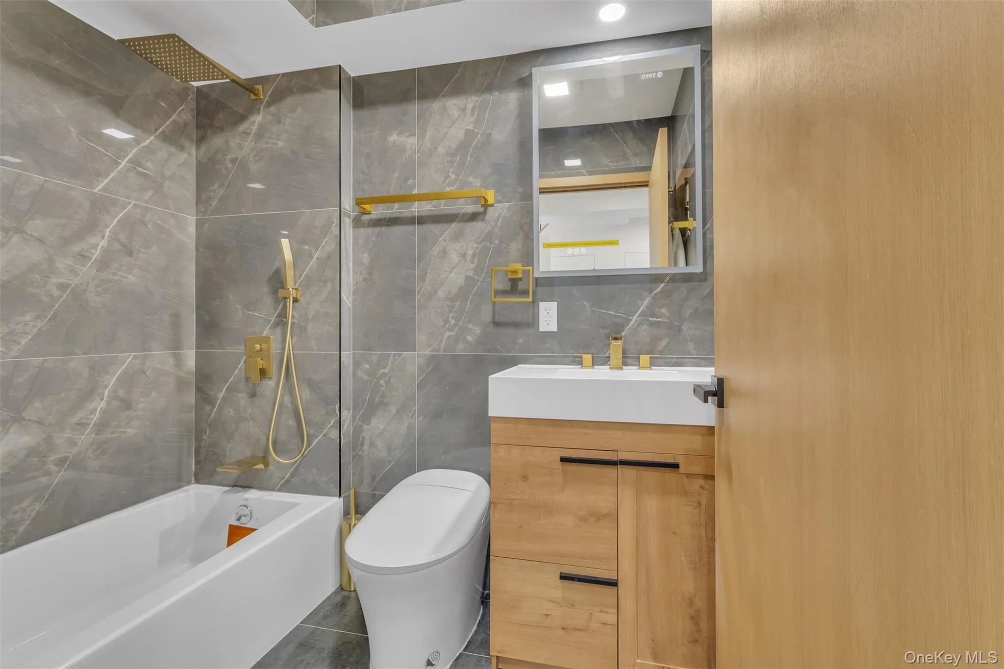 Bathroom featuring wall-to-ceiling large-format tile, vanity with wood-finish cabinetry, brass-tone plumbing fixtures, wall-mounted mirror, and a built-in bathtub with integrated rain showerhead Bathroom featuring wall-to-ceiling large-format tile, vanity with wood-finish cabinetry, brass-tone plumbing fixtures, wall-mounted mirror, and a built-in bathtub with integrated rain showerhead