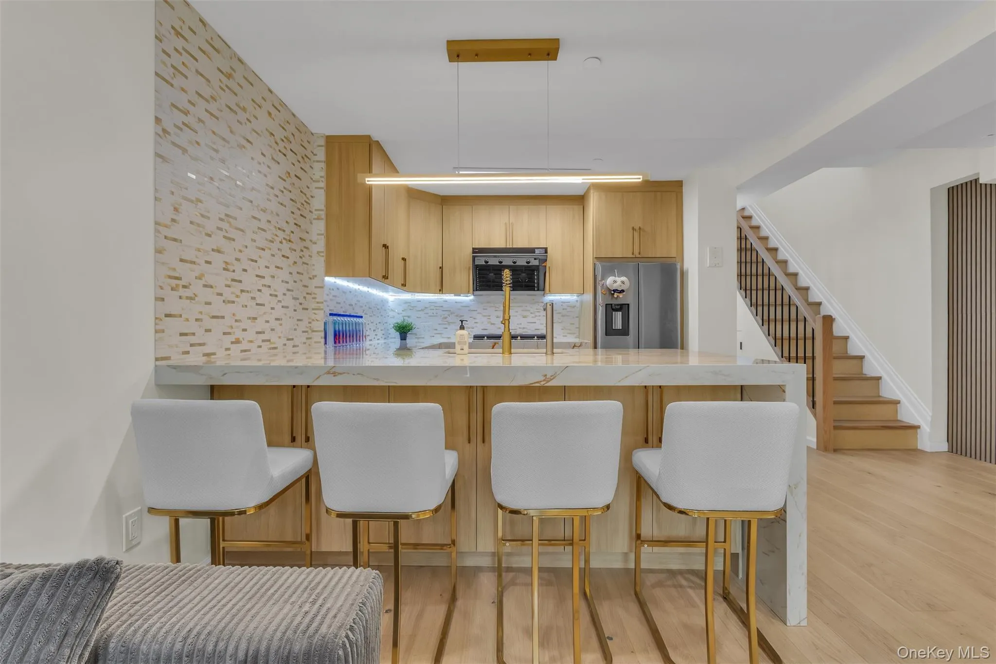 Kitchen featuring light wood-finish cabinetry, waterfall-edge stone countertop, decorative tile backsplash, integrated stainless steel appliances, and a linear pendant light fixture Kitchen featuring light wood-finish cabinetry, waterfall-edge stone countertop, decorative tile backsplash, integrated stainless steel appliances, and a linear pendant light fixture