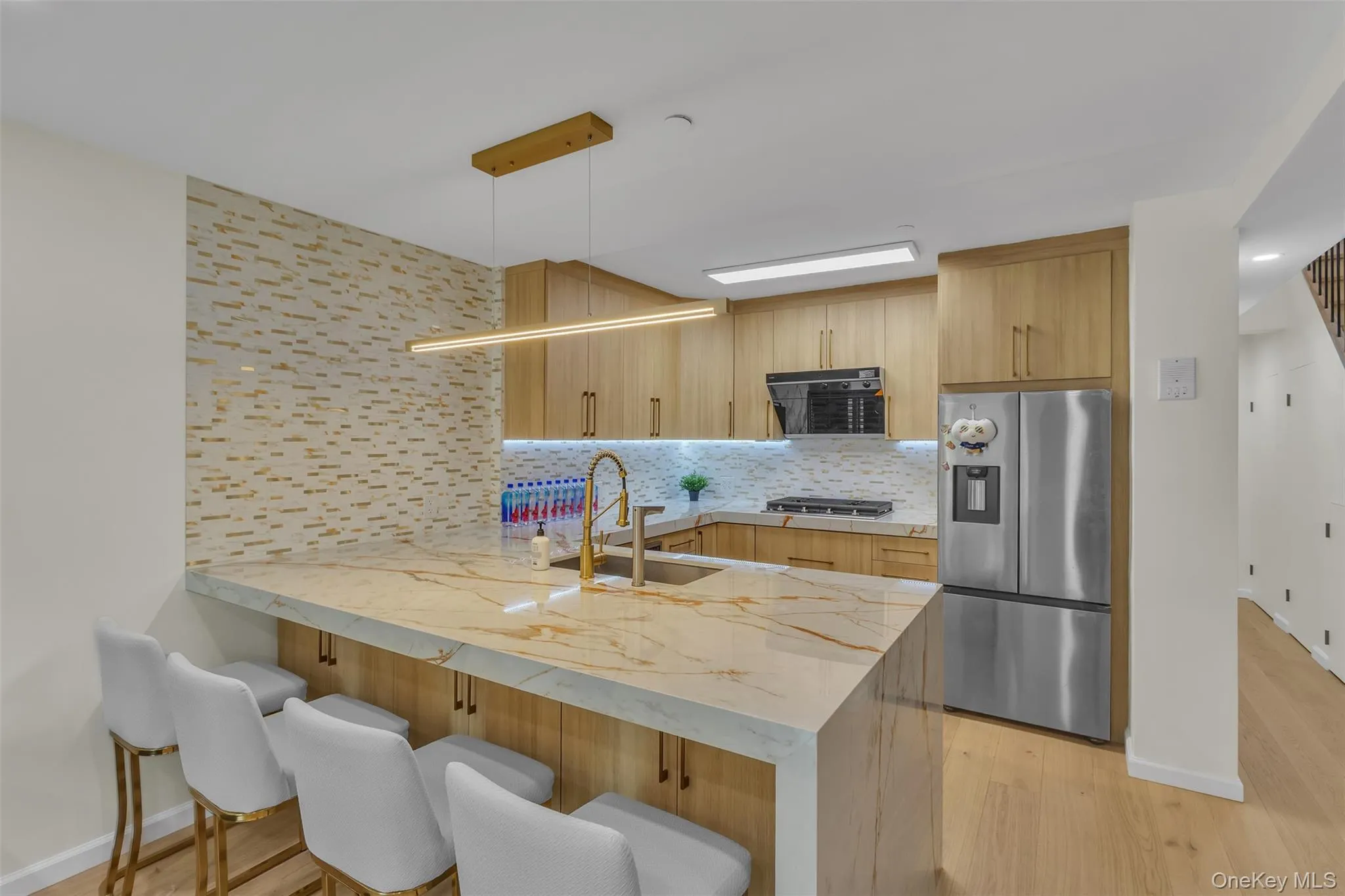 Kitchen featuring a waterfall-edge island with stone-finish countertops, wood-finish cabinetry, a tiled backsplash, wood-finish flooring, and a linear pendant light fixture Kitchen featuring a waterfall-edge island with stone-finish countertops, wood-finish cabinetry, a tiled backsplash, wood-finish flooring, and a linear pendant light fixture