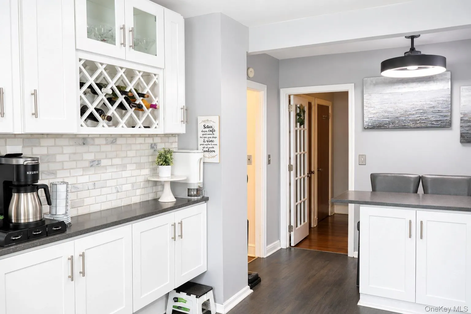Kitchen featuring white cabinetry with integrated wine rack, stone-tiled backsplash, dark solid-surface countertops, wood-finish flooring, and a modern drum-style pendant light fixture Kitchen featuring white cabinetry with integrated wine rack, stone-tiled backsplash, dark solid-surface countertops, wood-finish flooring, and a modern drum-style pendant light fixture