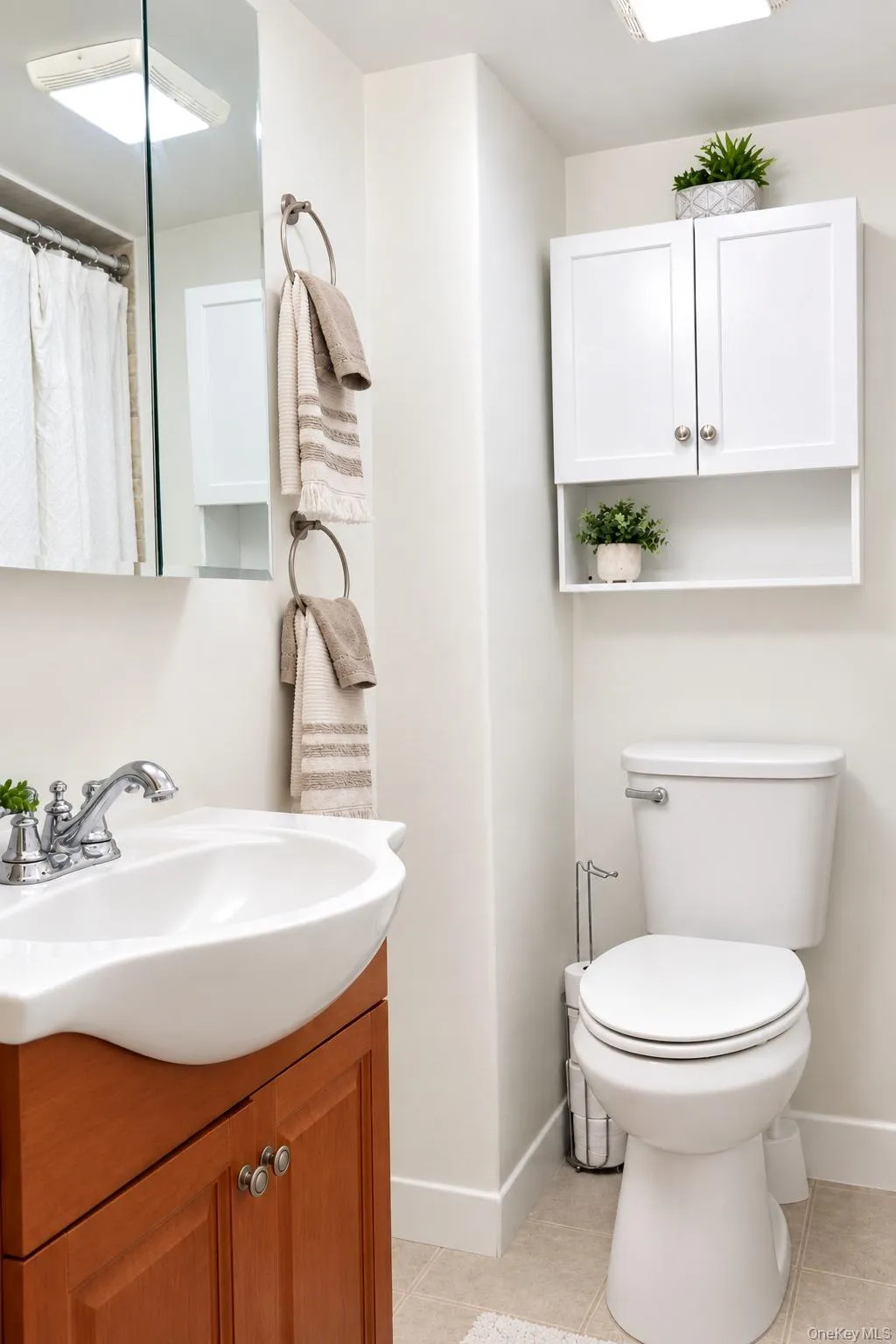 Wall-mounted white cabinetry with open shelving, wood-finish vanity, polished chrome faucet, rectangular wall-mounted mirror, and standard-height toilet Wall-mounted white cabinetry with open shelving, wood-finish vanity, polished chrome faucet, rectangular wall-mounted mirror, and standard-height toilet